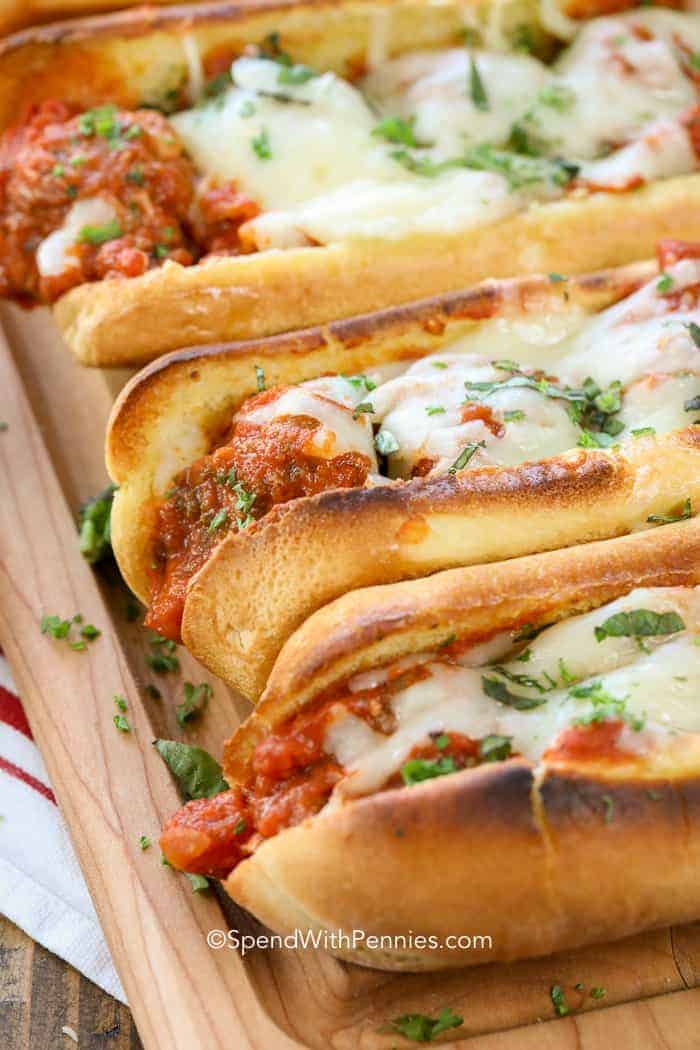 Three meatball subs on a wooden board, topped with melted cheese, marinara sauce, and chopped herbs—perfect for hearty Sports Dinners.