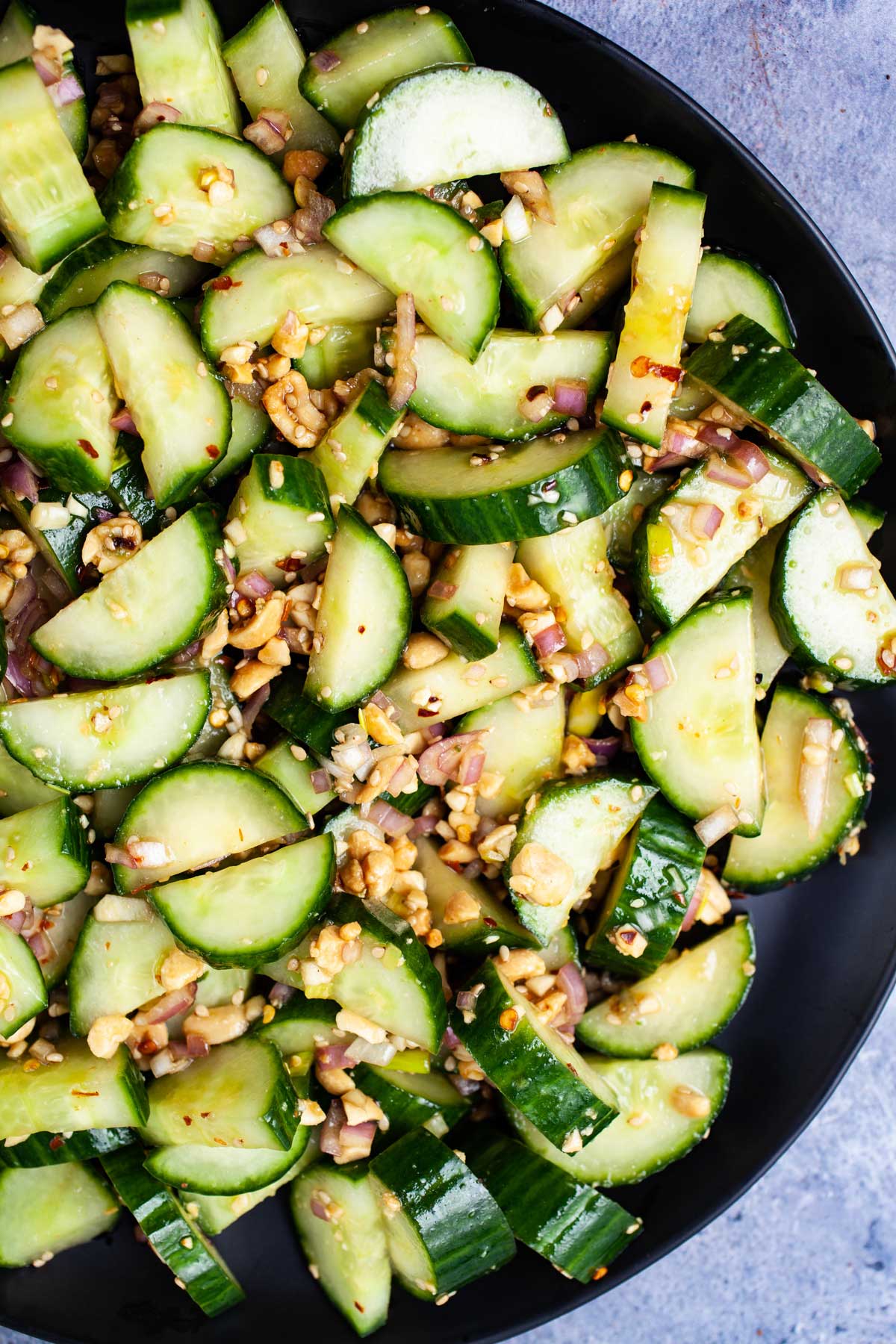 Sliced cucumbers mixed with chopped nuts, red onions, and seasonings on a black plate—an easy addition to your summer vegetable side dishes.