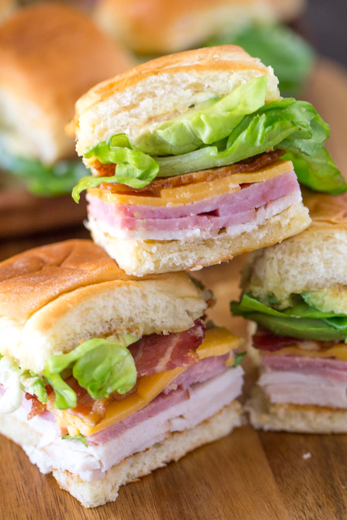 Small sandwiches with layers of ham, turkey, cheddar cheese, bacon, and lettuce on soft buns—perfect beach meals arranged on a wooden surface.
