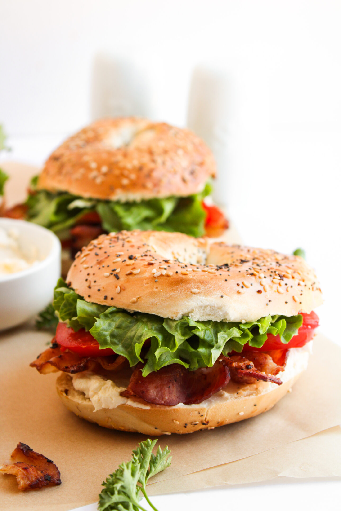 Two everything bagels filled with bacon, lettuce, and tomato sit on parchment paper, with a bowl of sauce and herbs in the background—perfect for easy summer meals or fresh June recipes.