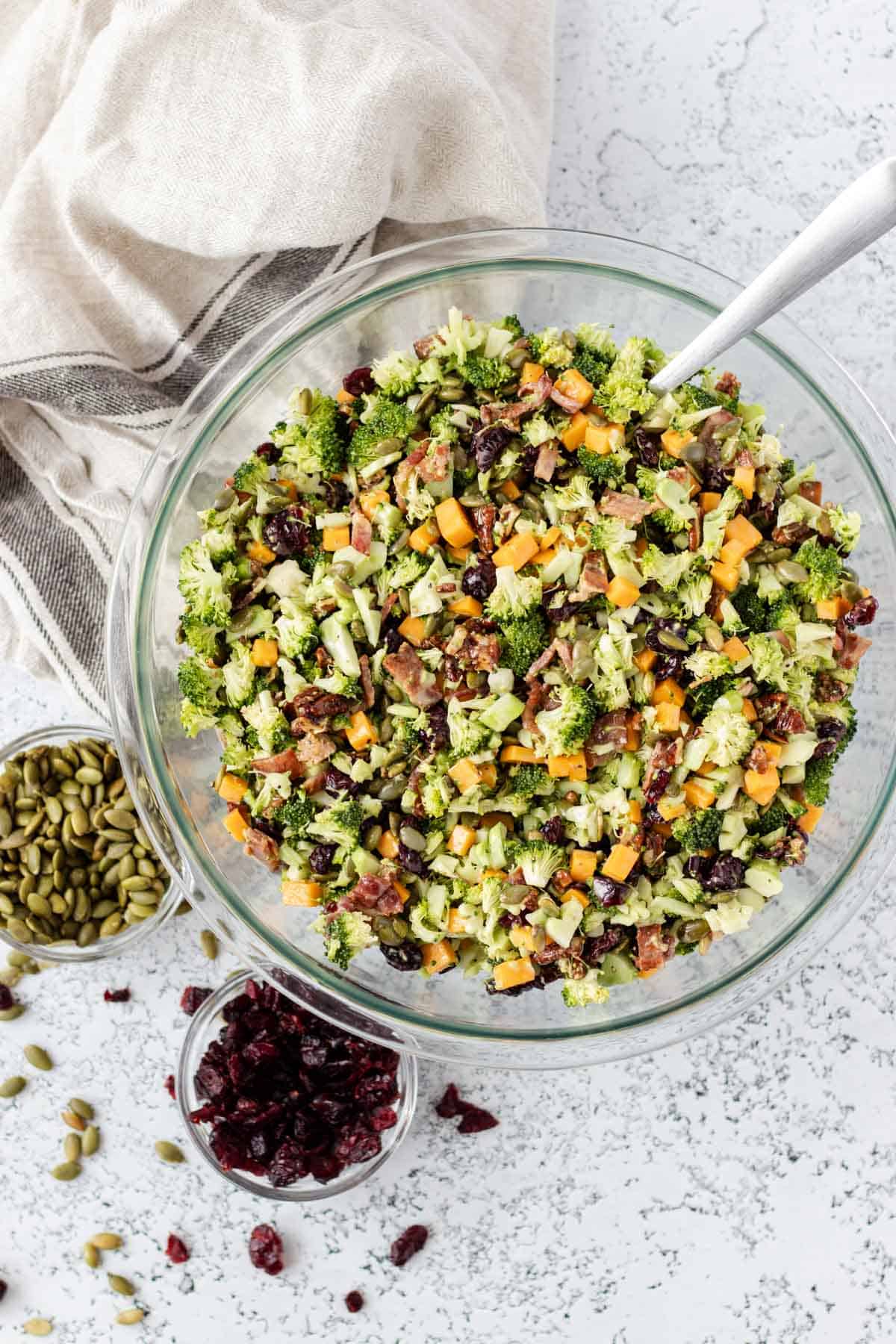 A glass bowl filled with chopped broccoli salad—one of the best summer vegetable side dishes—mixed with cheddar cheese, bacon, dried cranberries, and seeds. Small bowls of pumpkin seeds and cranberries sit beside it, ready to enjoy.