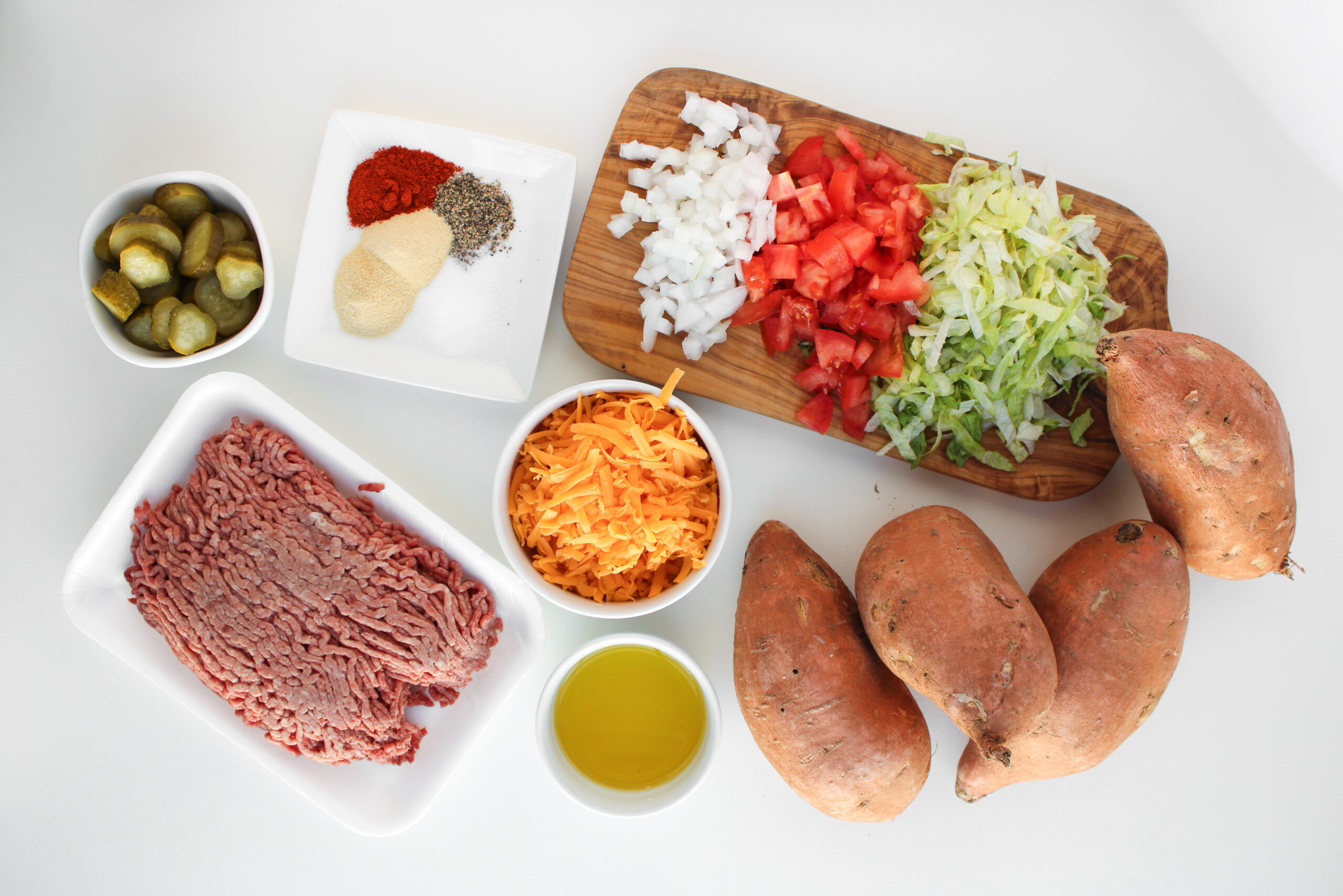 Ingredients for loaded sweet potatoes arranged on a white surface, featuring ground beef, shredded cheese, olive oil, sweet potatoes, diced onions, tomatoes, lettuce, pickles, and spices. A perfect twist on classic cheeseburger flavors.