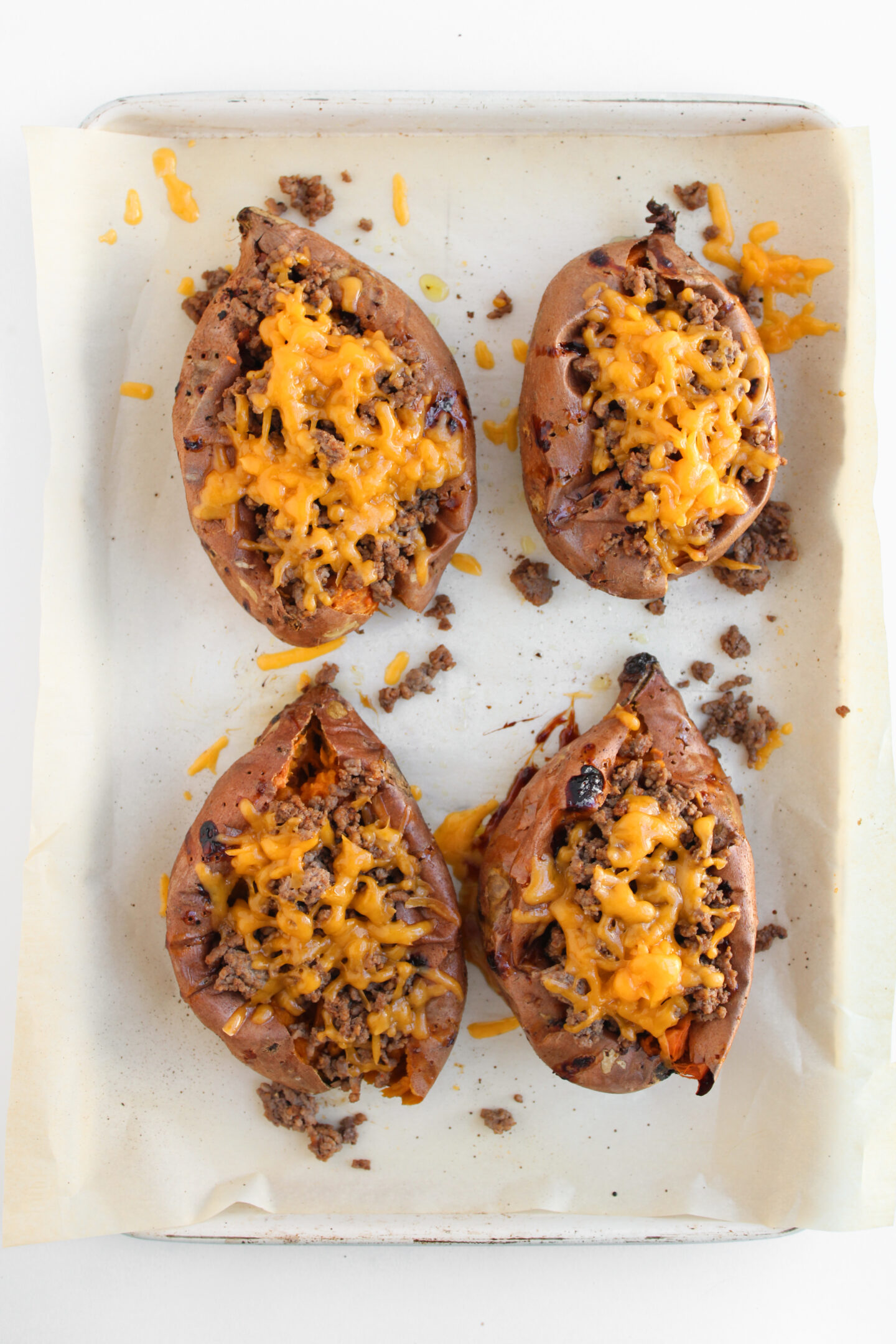 Four loaded sweet potatoes topped with savory ground beef and melted cheddar cheese, resembling a delicious cheeseburger, all on a parchment-lined baking sheet.
