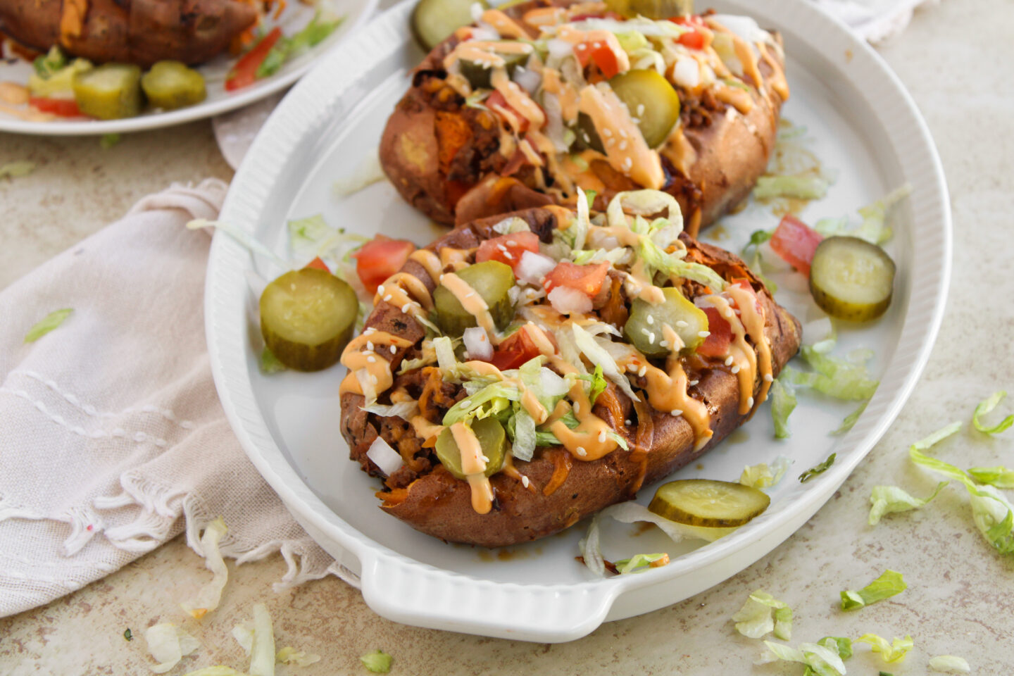 Cheeseburger stuffed sweet potatoes topped with shredded lettuce, diced tomato, onions, pickles, and a drizzle of creamy sauce, served on a white oval dish with pickle slices on the side—a delicious high protein dinner option.