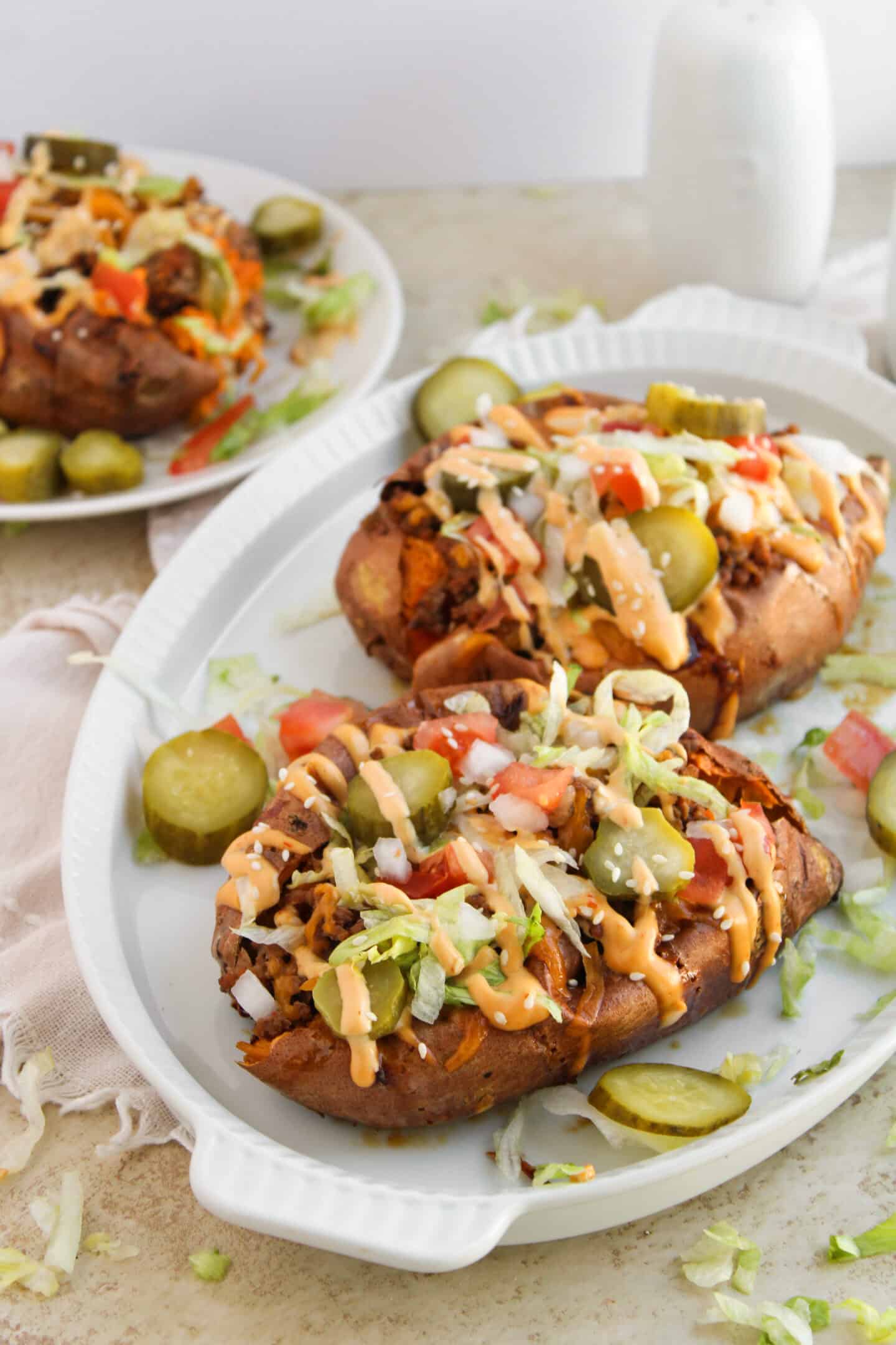 Two baked sweet potatoes are transformed into loaded sweet potatoes, topped with shredded lettuce, diced tomatoes, pickles, and a creamy sauce, served on a white oval dish with extra pickle slices—like a cheeseburger twist.