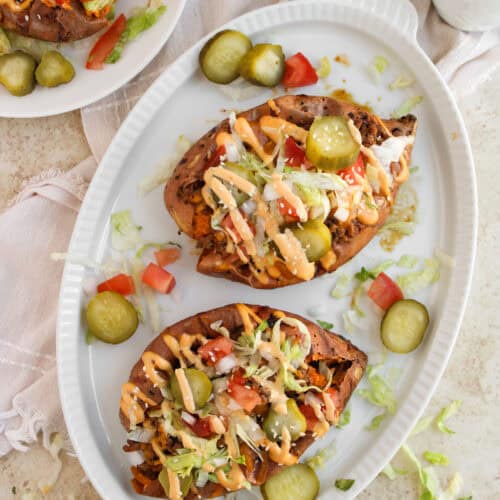 Cheeseburger loaded sweet potatoes are topped with ground meat, shredded lettuce, diced tomatoes, pickles, and sauce on an oval white plate, with extra pickle slices on the side.