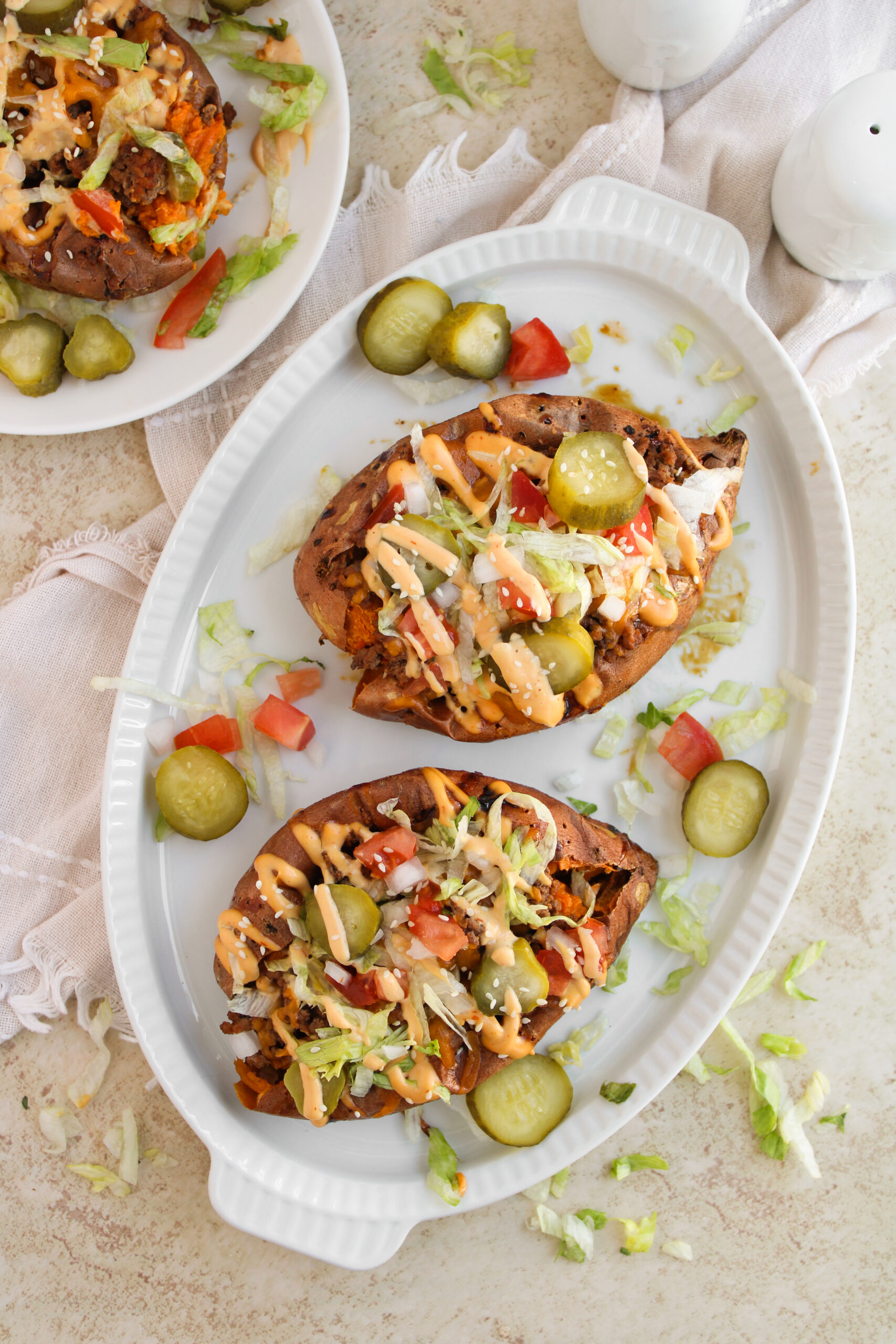 Cheeseburger loaded sweet potatoes are topped with ground meat, shredded lettuce, diced tomatoes, pickles, and sauce on an oval white plate, with extra pickle slices on the side.
