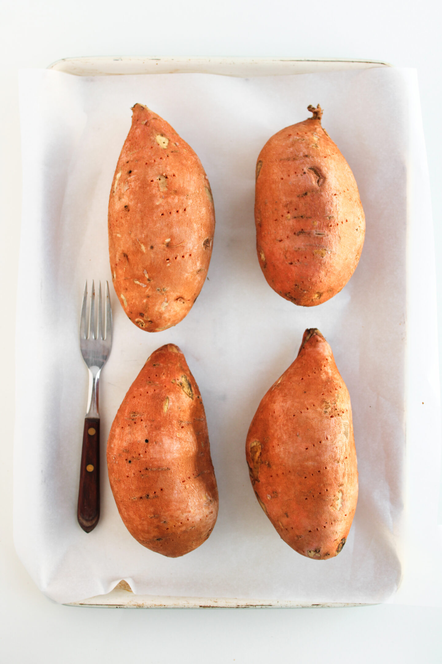 Four whole sweet potatoes and a fork are arranged on a parchment-lined baking sheet, ready to be transformed into delicious loaded sweet potatoes—one of the best sweet potato recipes to try.