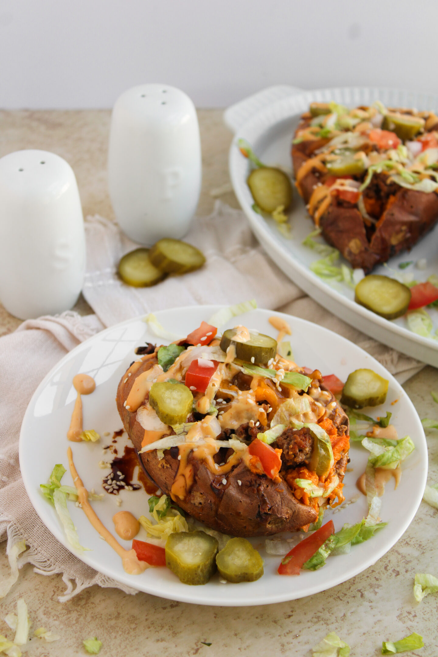 A baked sweet potato on a plate topped with sliced pickles, lettuce, tomatoes, shredded cheese, and sauce—perfect for summer meals or creative dinner ideas—with salt and pepper shakers in the background.
