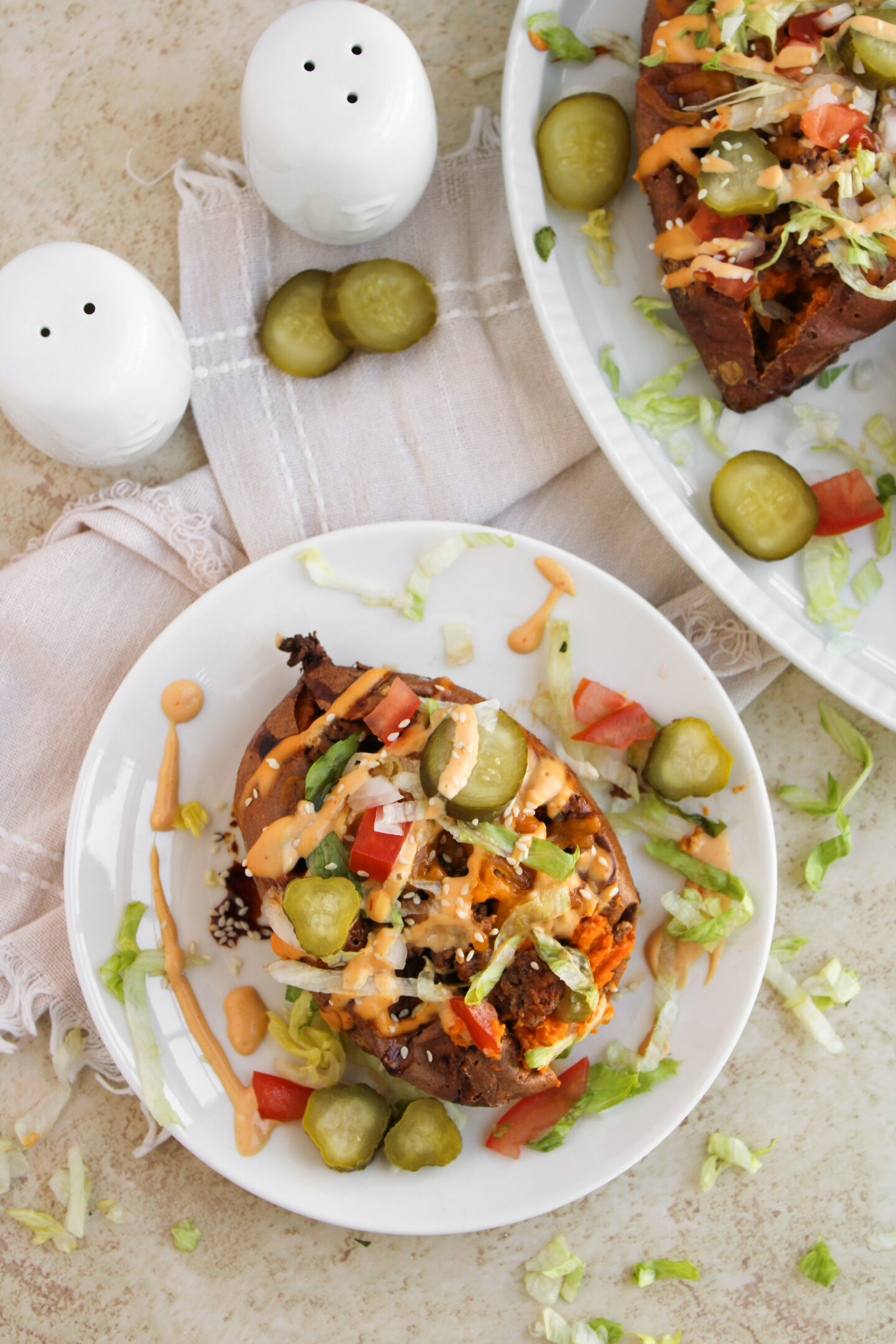 A baked potato topped with ground beef, shredded lettuce, sliced pickles, diced tomatoes, and a creamy sauce makes a hearty addition to your summer recipes—served on a white plate next to salt and pepper shakers.