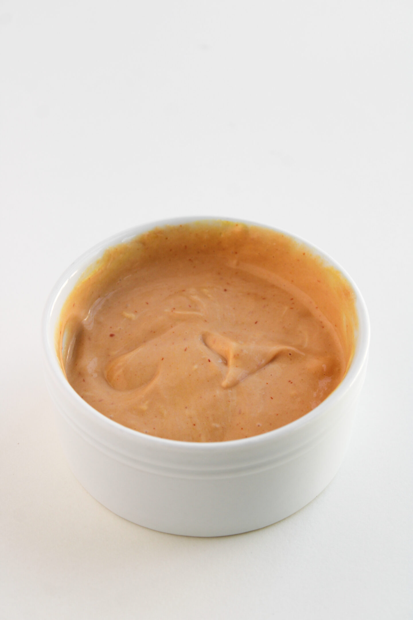 A small white bowl filled with a creamy, light orange dipping sauce—perfect for pairing with loaded sweet potatoes—sits on a white surface.