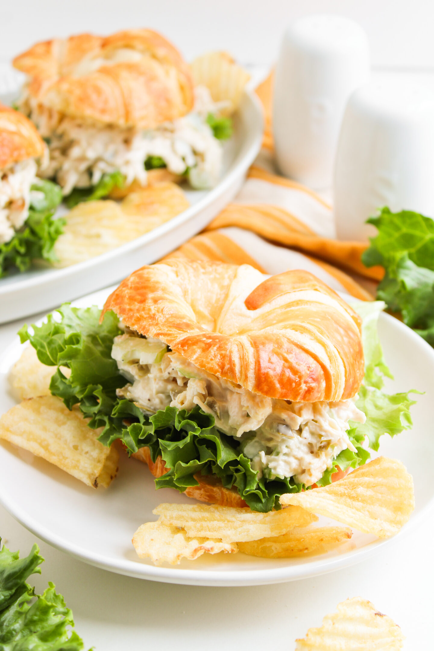 A croissant sandwich with lettuce and chicken salad is served on a white plate with potato chips—perfect for casual Sports Dinners. A plate with another sandwich and salt and pepper shakers are in the background.