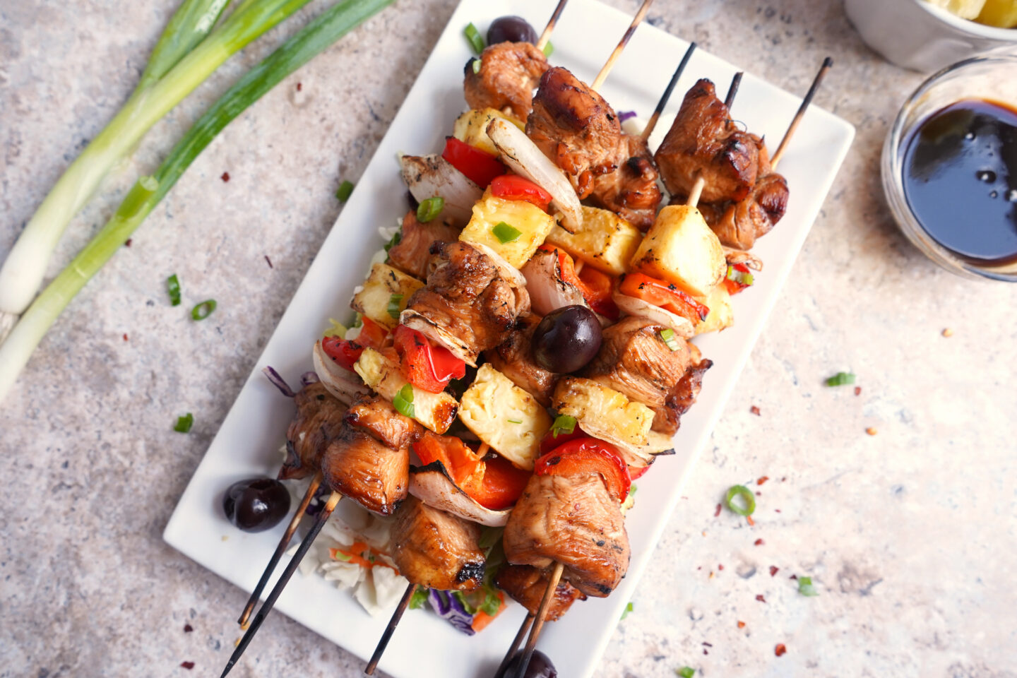Five grilled kebab skewers with chicken, pineapple, onions, peppers, and tomatoes are arranged on a rectangular white plate with olives and a bowl of sauce nearby—perfect for Father's Day dinner or festive dinner ideas.