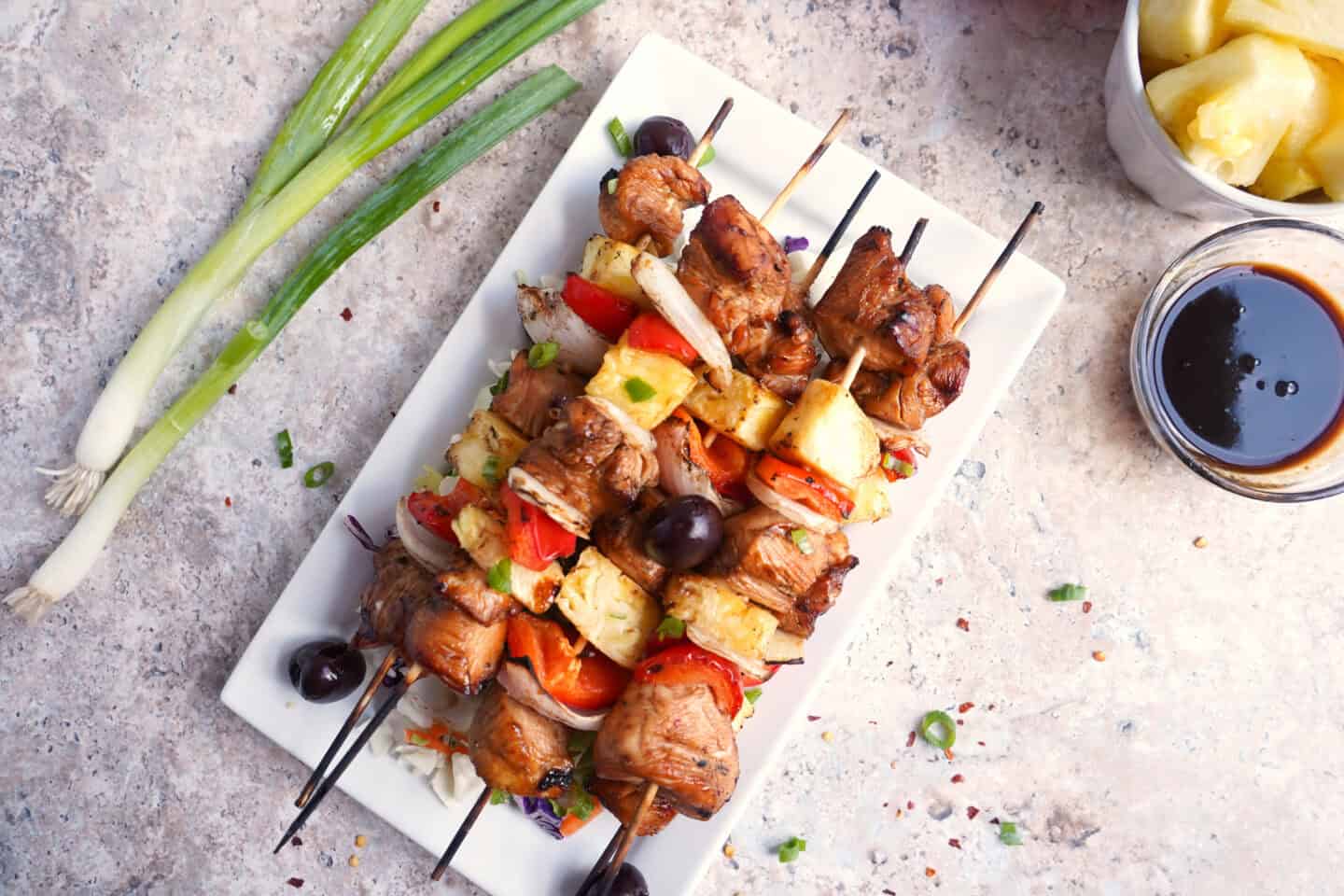 Grilled chicken and vegetable skewers served on a white rectangular plate, with green onions, a bowl of pineapple, and a small bowl of sauce on the side—perfect for beach dinner or easy summer dinner ideas.