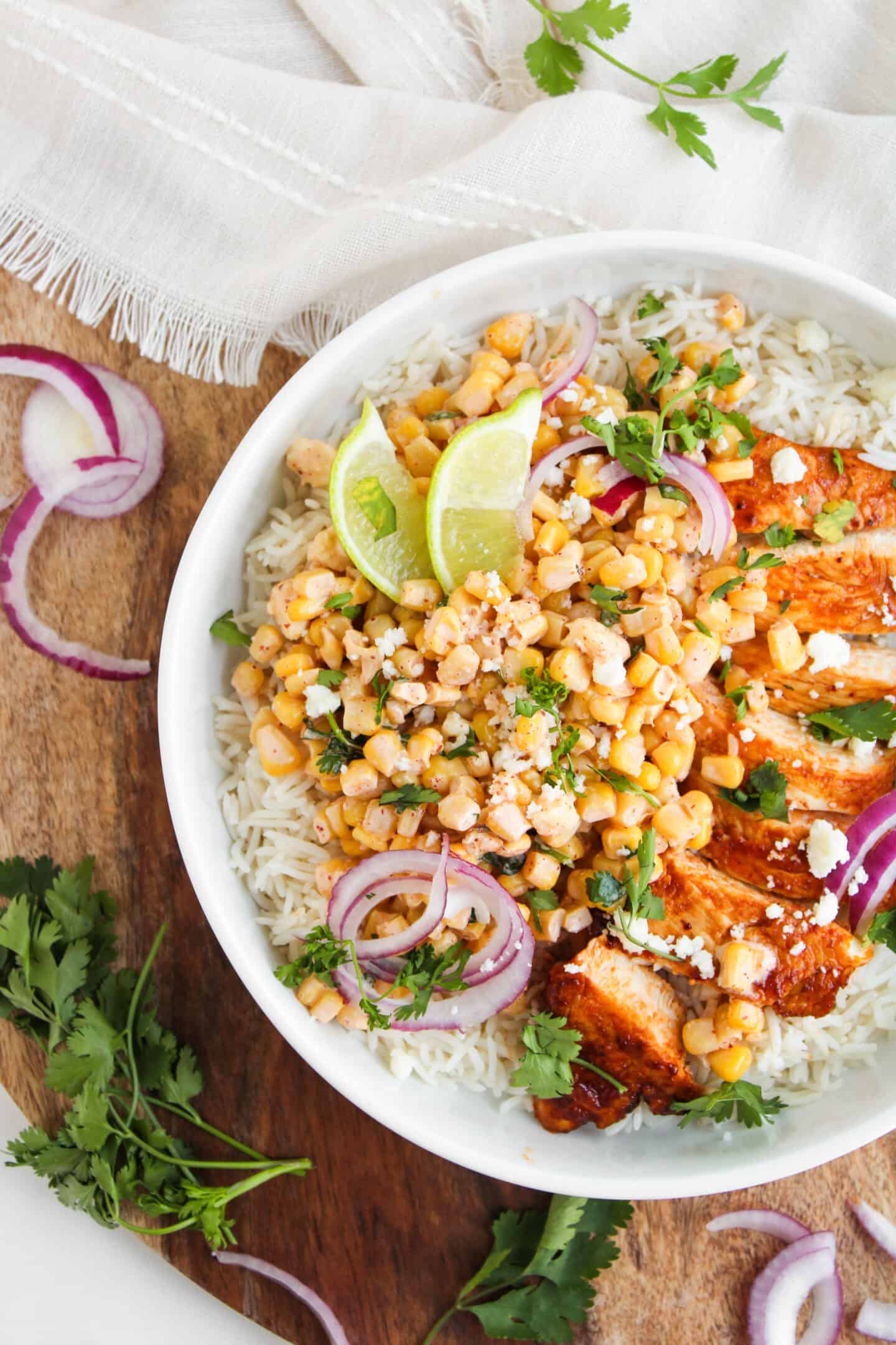 A bowl of rice topped with grilled chicken, corn, chickpeas, red onion, cilantro, lime wedges, and crumbled cheese—one of the best easy summer recipes for fresh summer dinners. Served on a wooden surface with fresh herbs nearby.