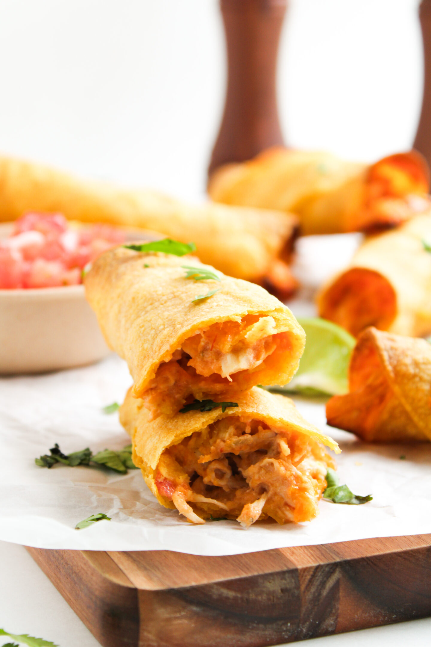 Two crispy, golden-brown taquitos filled with shredded chicken are stacked on a wooden board—perfect for casual Sports Dinners—with a bowl of salsa and other taquitos in the background.