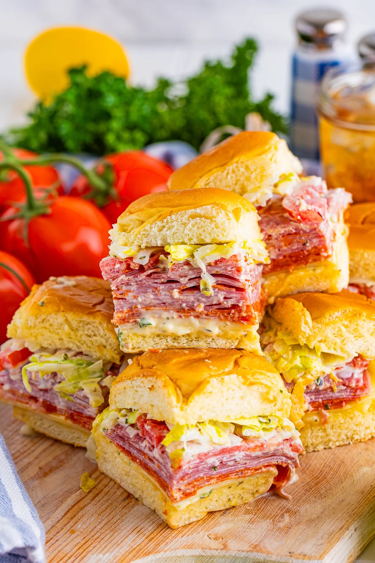 A stack of deli meat and cheese sliders with lettuce on soft buns, perfect for Sports Dinners, displayed on a wooden board with fresh tomatoes and parsley in the background.
