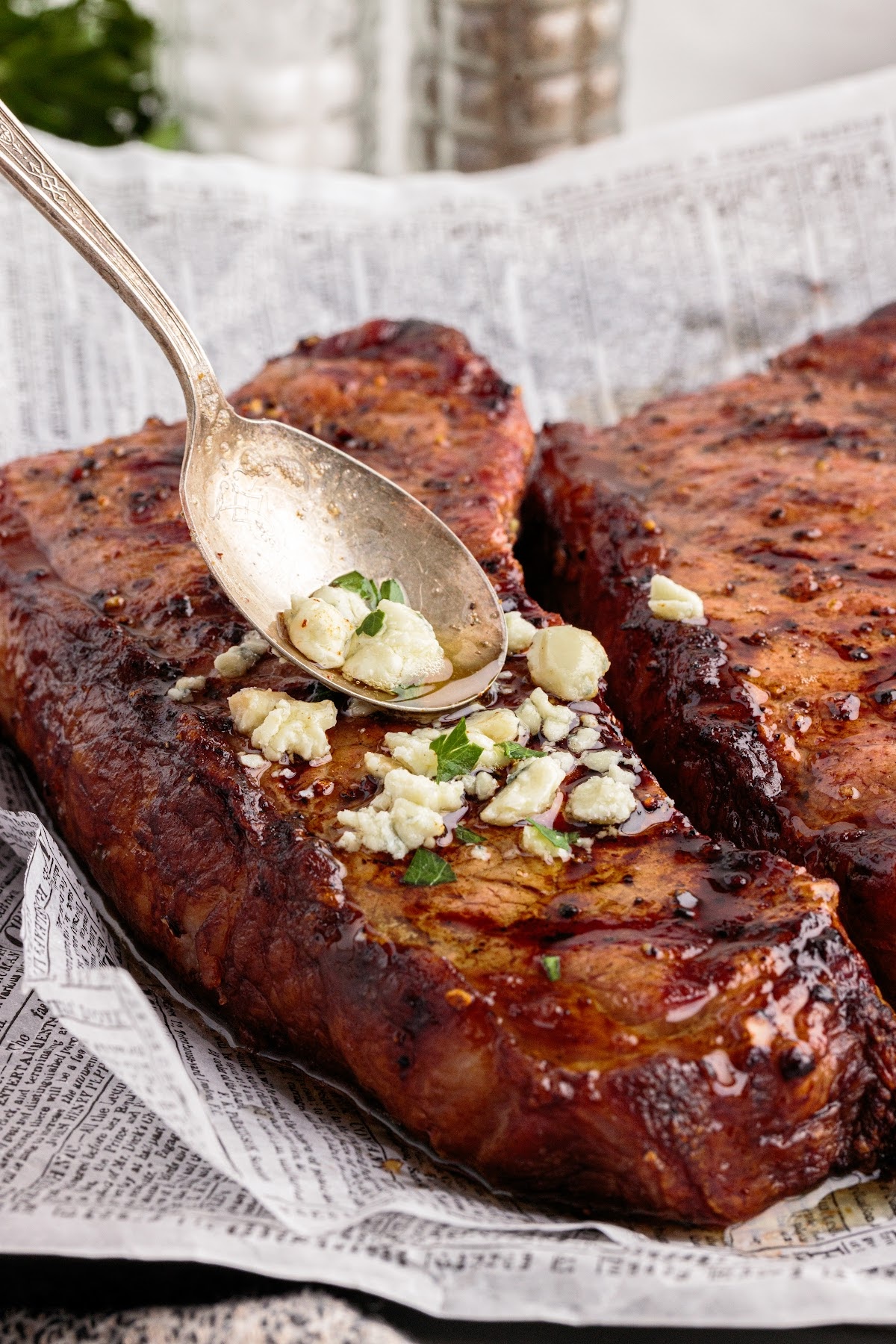A spoon adds blue cheese crumbles and herbs to two grilled steaks resting on newspaper—perfect inspiration for unique Father's Day dinner ideas.