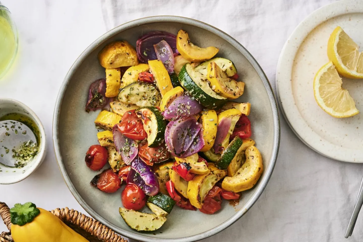 A bowl of roasted vegetables—zucchini, yellow squash, red onion, and cherry tomatoes—garnished with herbs, makes a vibrant addition to summer vegetable side dishes. A plate with lemon wedges sits beside it.