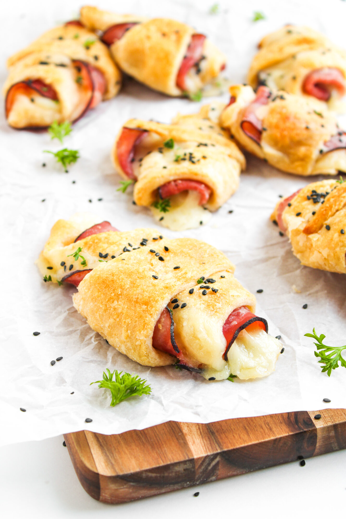Several ham and cheese crescent rolls topped with black sesame seeds are arranged on parchment paper, garnished with small sprigs of parsley—perfect as a tasty addition to your June Recipes or creative summer meals.