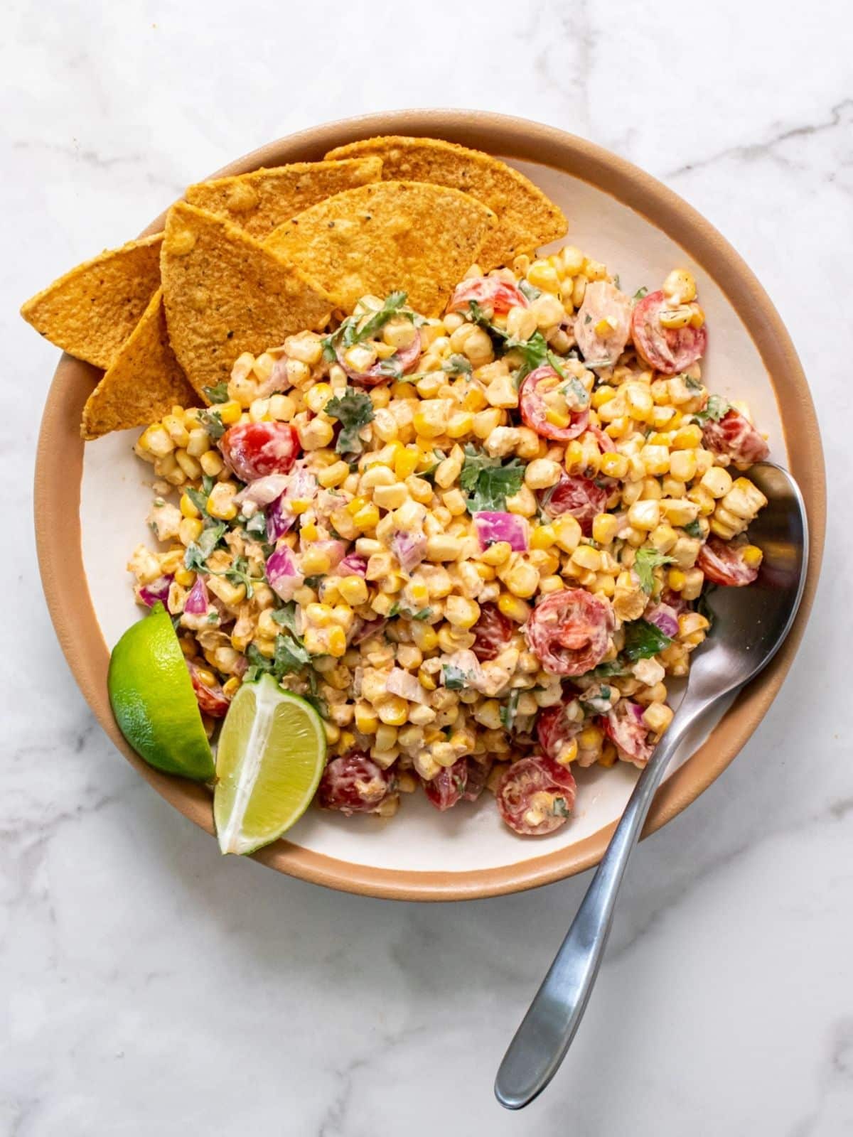 A plate of corn salad with chopped summer vegetables, garnished with lime wedges and served with tortilla chips, on a white marble surface with a spoon—a fresh pick for summer vegetable side dishes.
