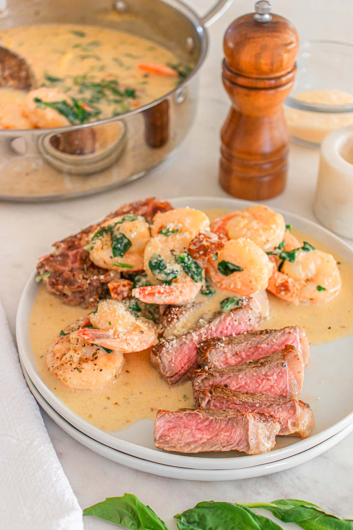 A plate of sliced steak topped with shrimp in a creamy sauce, garnished with herbs—an irresistible Father’s Day dinner idea, with a skillet of the same dish and a pepper grinder in the background.
