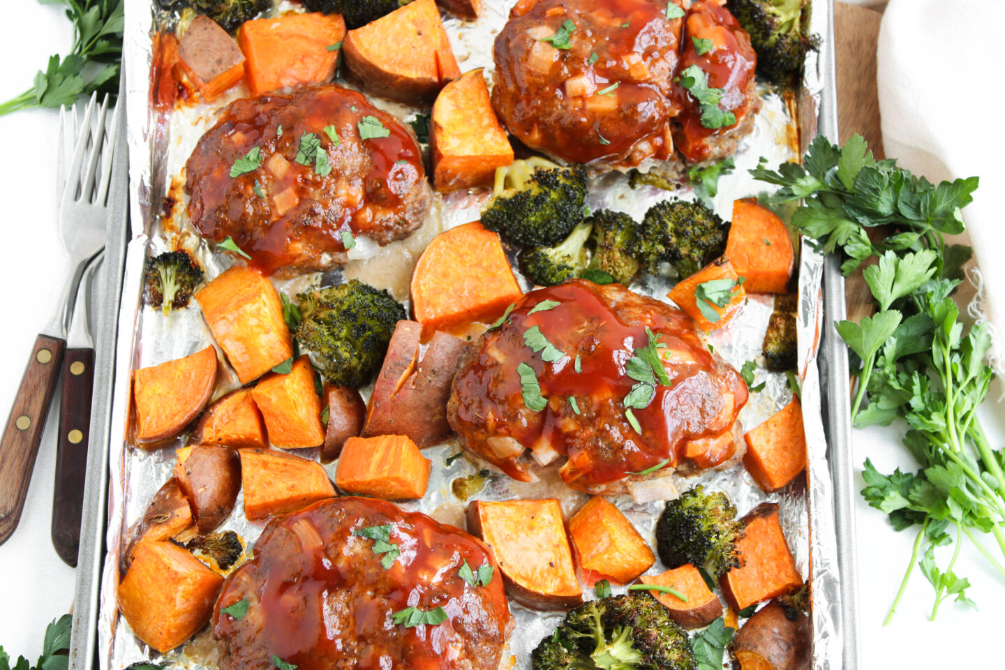 A sheet pan with four glazed mini meatloafs, roasted sweet potato chunks, and broccoli, garnished with chopped herbs—perfect for Father's Day dinner ideas.