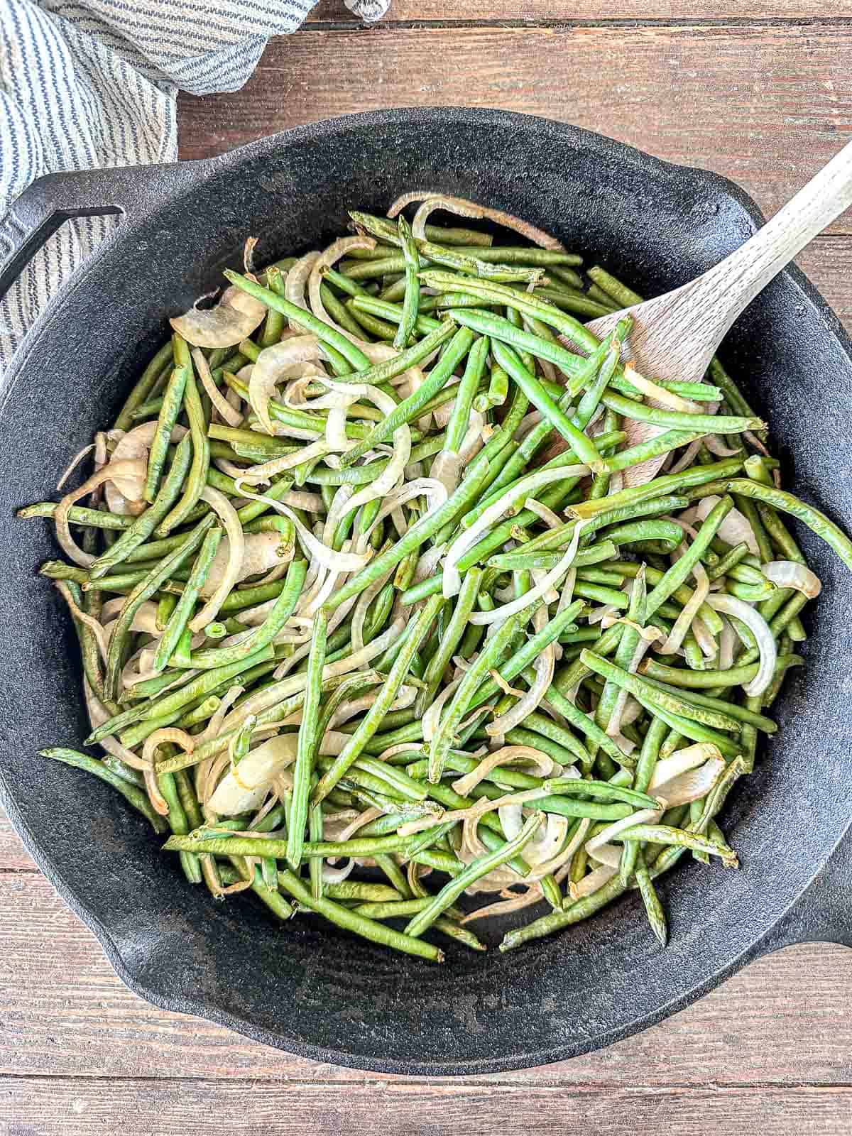 Sautéed green beans and sliced onions in a black cast iron skillet make a perfect summer vegetable side dish, stirred with a wooden spoon on a wooden surface.