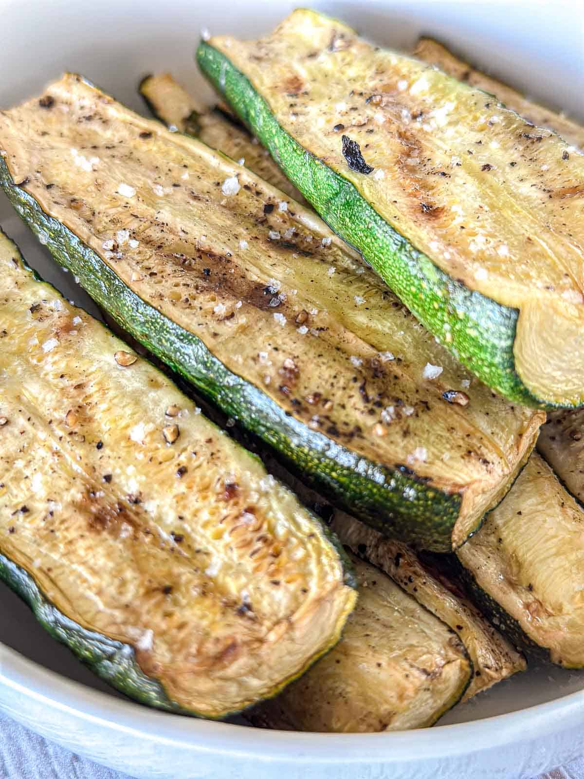 Roasted zucchini spears, a perfect addition to summer vegetable side dishes, are seasoned with salt and black pepper and arranged in a white bowl.