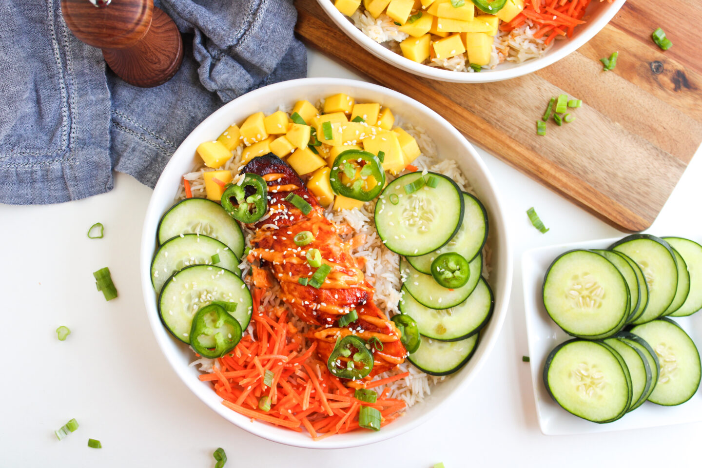 A bowl filled with rice, sliced grilled chicken, shredded carrots, mango cubes, cucumber slices, jalapeños, and green onions—perfect for June recipes or a fresh summer dinner. Extra cucumber slices are served on a plate nearby.