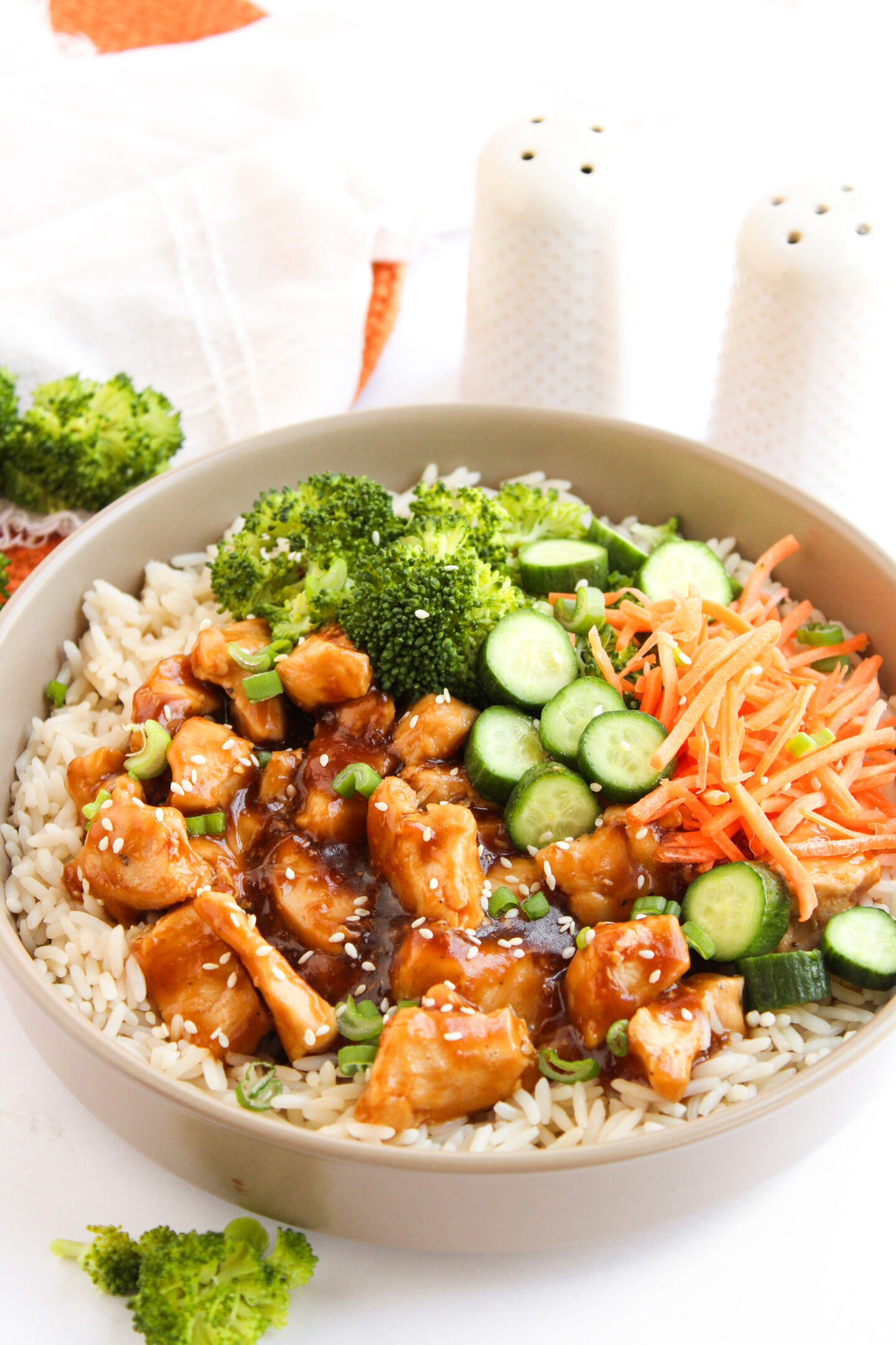 A bowl of rice topped with glazed chicken pieces, broccoli florets, sliced cucumber, and shredded carrots—perfect for summer meals or fresh June recipes—with salt and pepper shakers in the background.