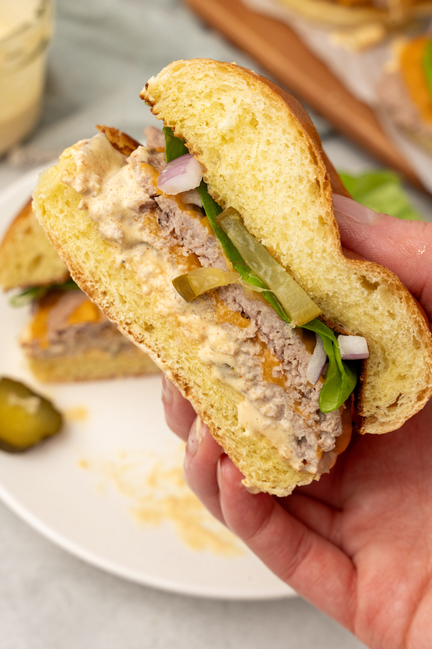 A hand holds half of a cheeseburger with pickles, lettuce, onions, and sauce on a brioche bun—perfect for beach meals—with the other half resting on a white plate in the background.