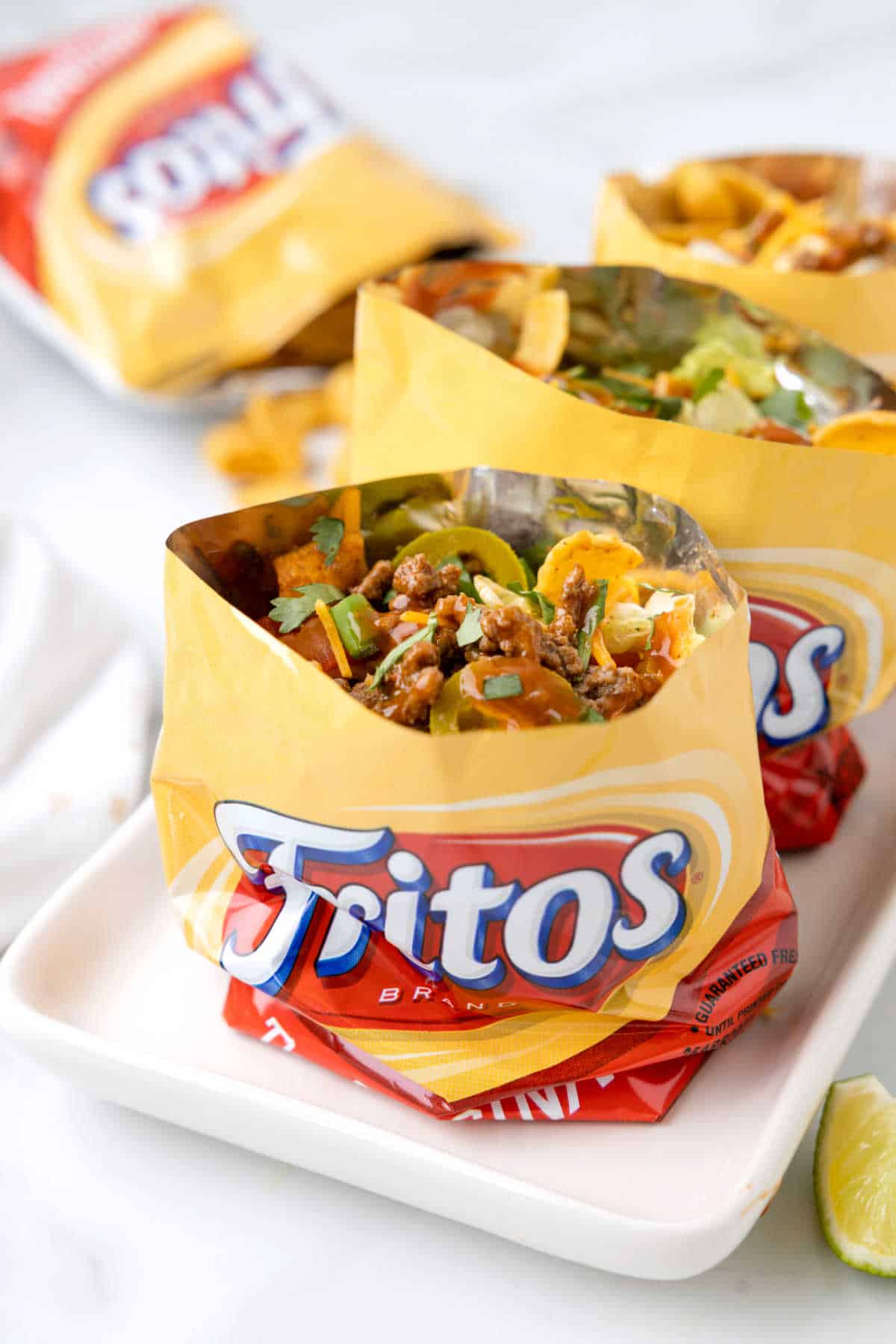Three small bags of Fritos are opened and filled with chili, cheese, green onions, and cilantro—an easy beach food perfect for fun dinner ideas, all served upright on a white tray.
