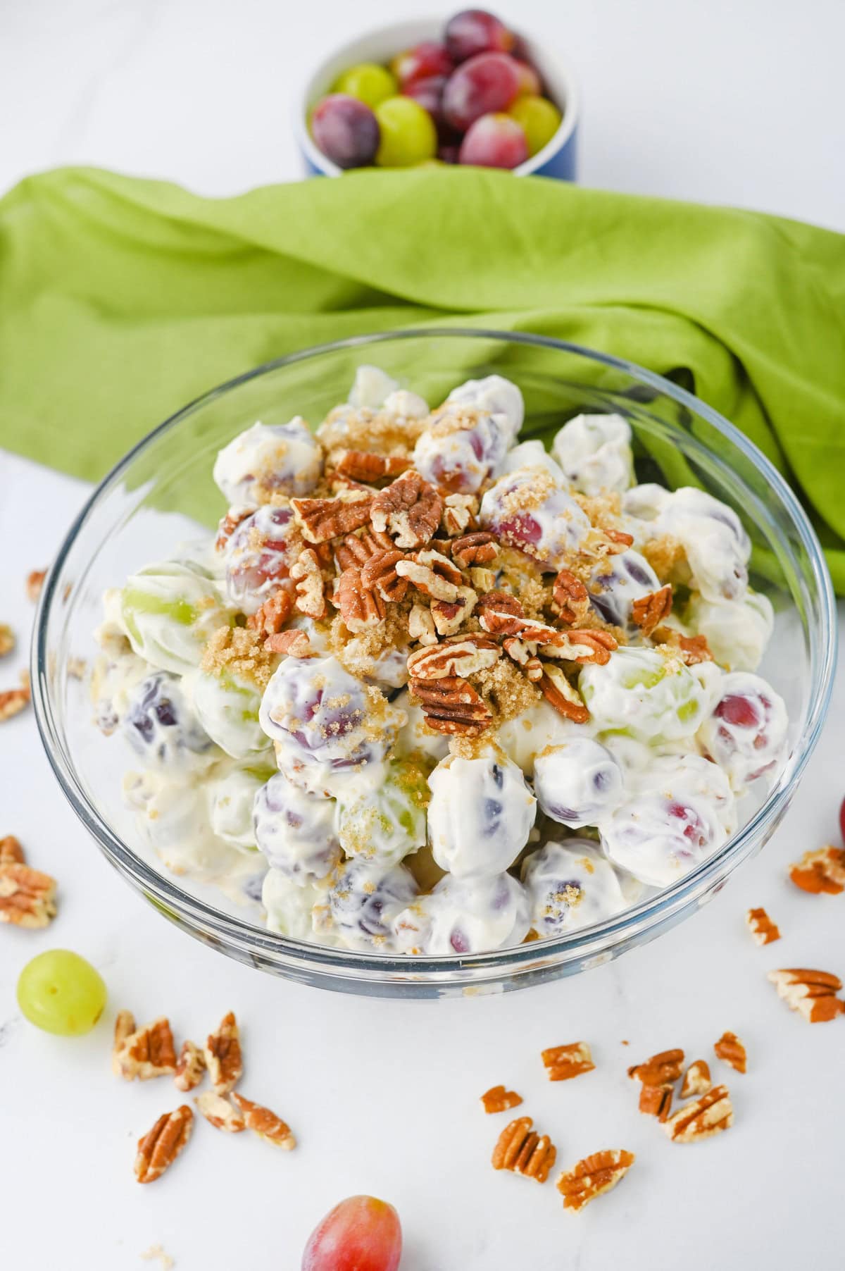 A glass bowl filled with creamy grape salad topped with chopped pecans, perfect for side dishes at a graduation party. Loose grapes and pecans are scattered on a white surface, with a small bowl of grapes in the background.