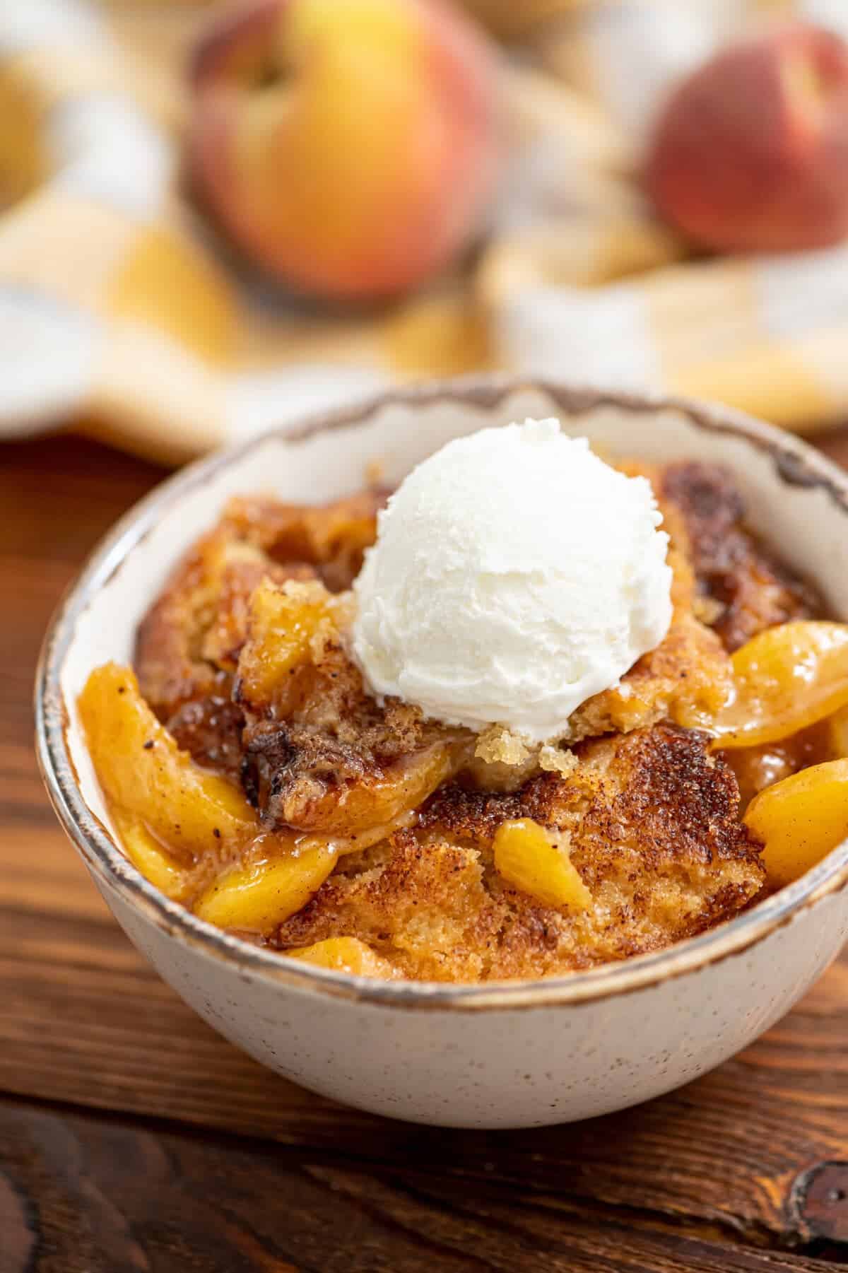 A bowl of peach cobbler, the perfect peach dessert, is topped with a scoop of vanilla ice cream, with fresh peaches in the background.