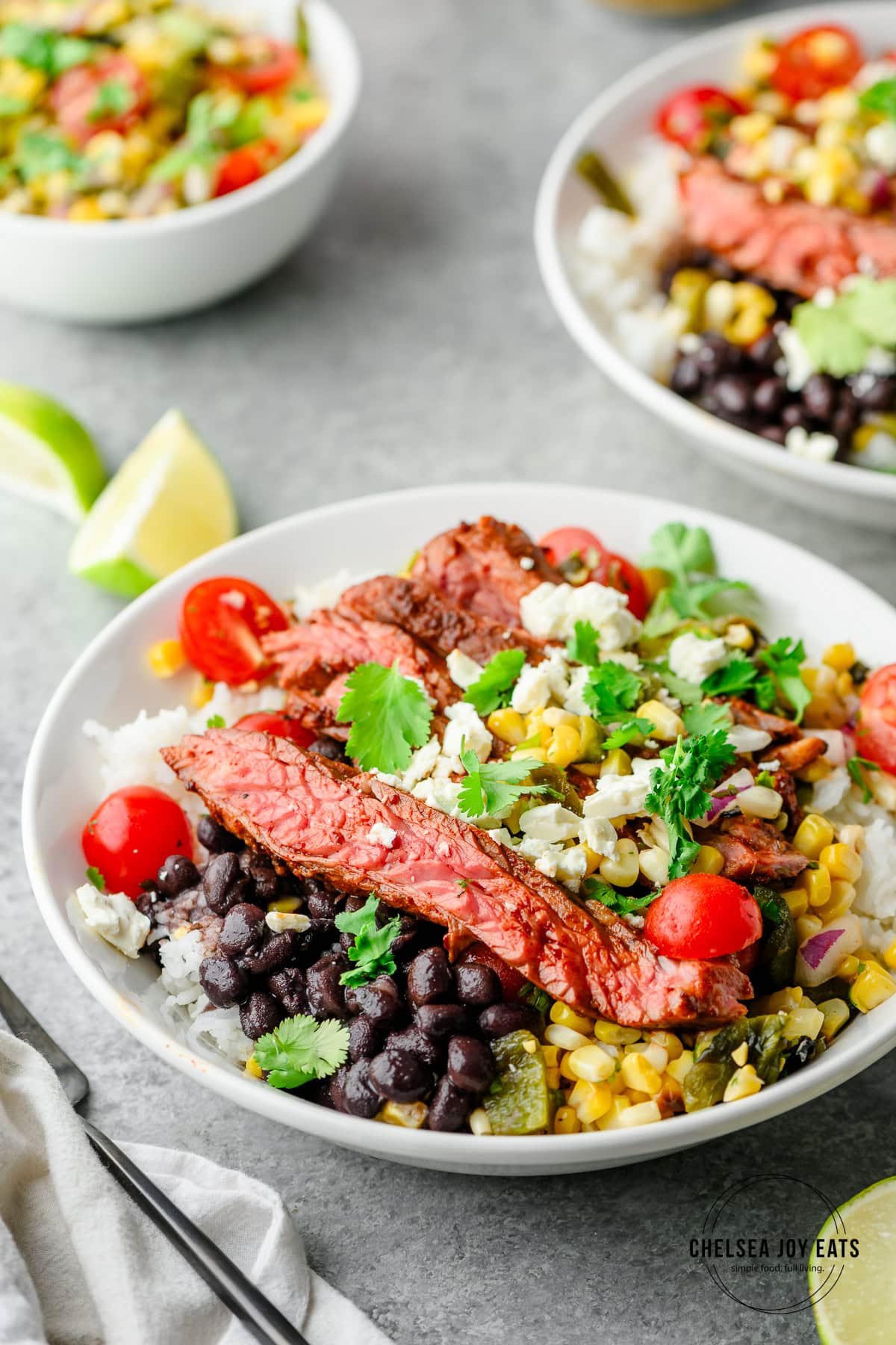 A high protein bowl of sliced steak, black beans, rice, corn, cherry tomatoes, cilantro, and cheese with refreshing lime wedges on the side—perfect for a summer meal.