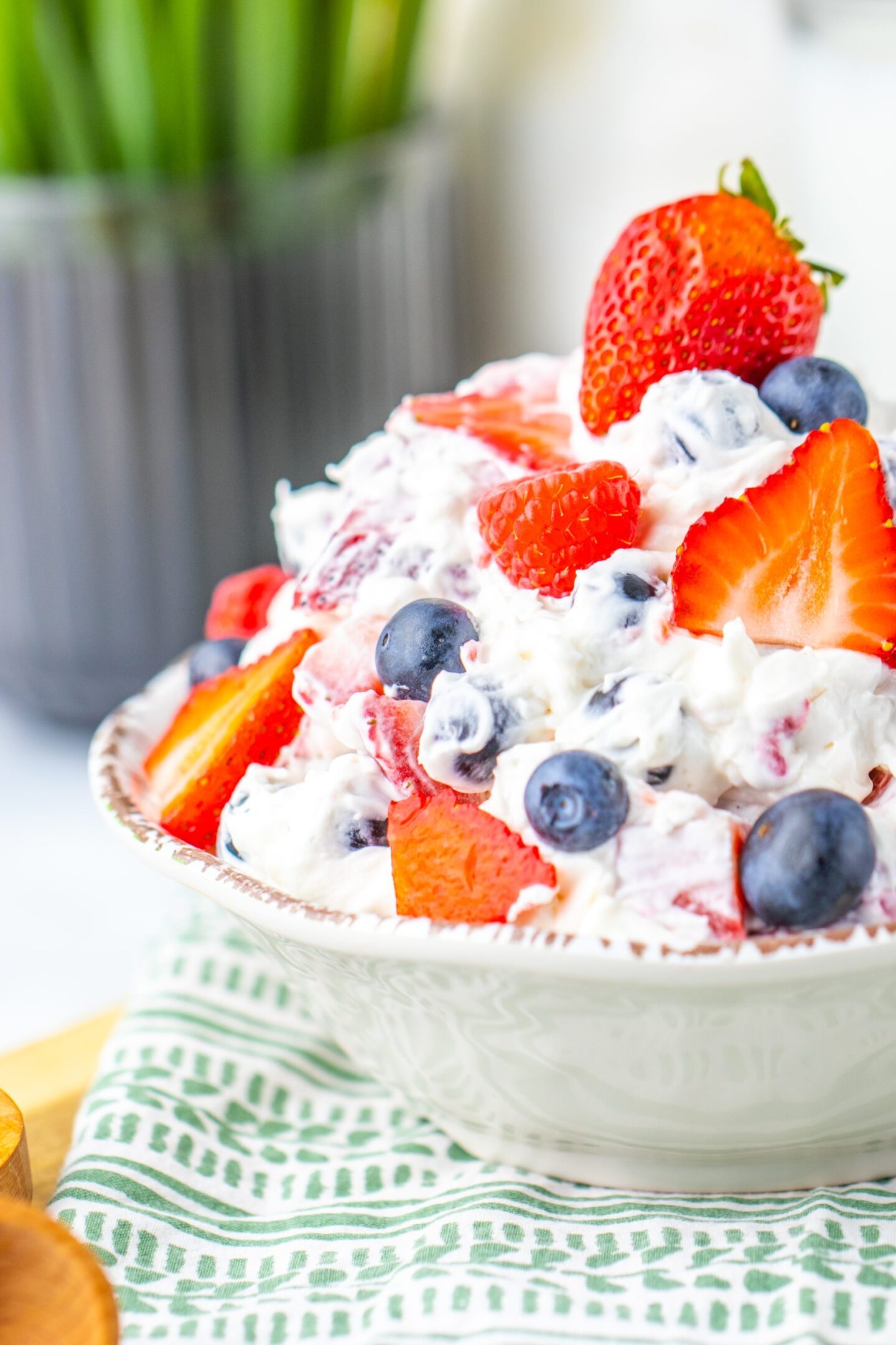 A bowl of whipped cream mixed with fresh strawberries and blueberries, ideal as party food, placed on a patterned cloth with a blurred plant in the background.