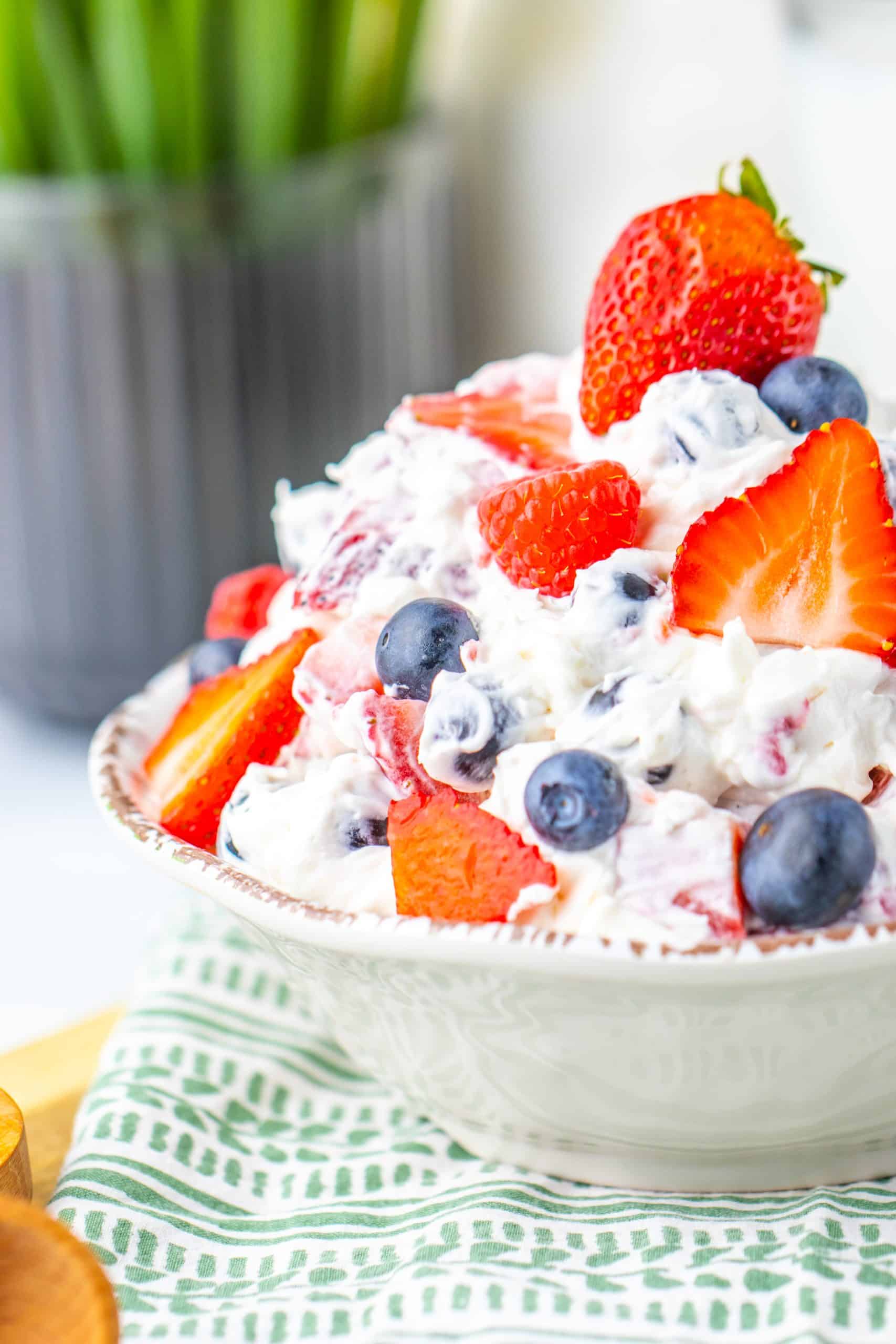 A bowl of whipped cream mixed with fresh strawberries and blueberries, ideal as party food, placed on a patterned cloth with a blurred plant in the background.