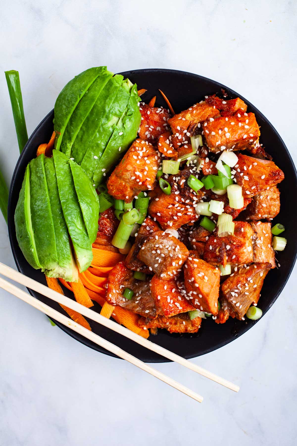 A black bowl filled with high protein tofu pieces topped with sesame seeds and green onions, sliced avocado, carrot ribbons, and chopsticks on a light surface—perfect for a fresh summer meal.