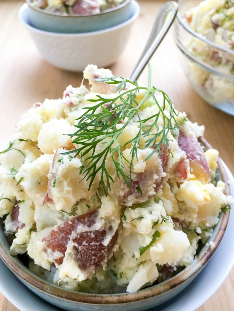 A bowl of potato salad with red-skinned potatoes, chopped herbs, and a sprig of dill on top, served with a spoon—perfect for side dishes at your graduation party.