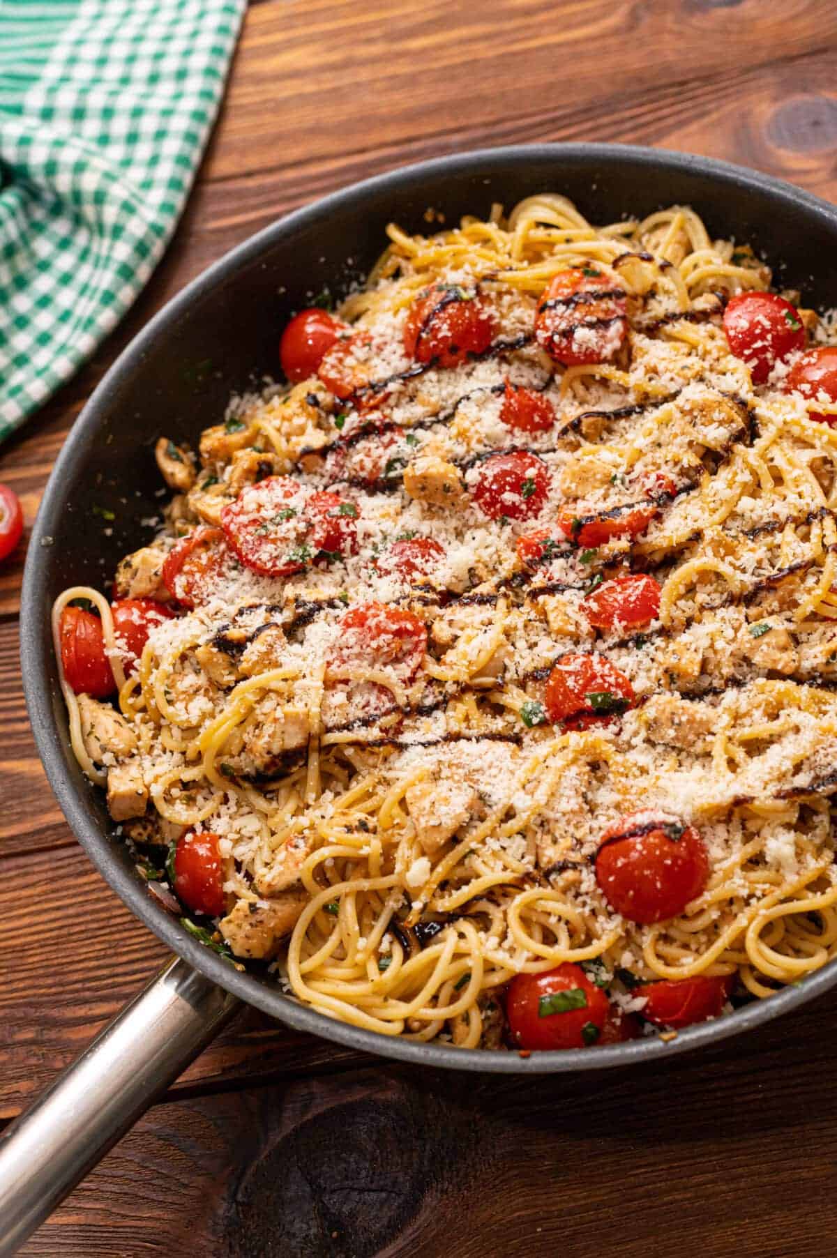 A skillet filled with spaghetti, cherry tomatoes, diced chicken, herbs, and grated cheese on a wooden table with a green checkered cloth nearby—perfect for Italian Summer Recipes.