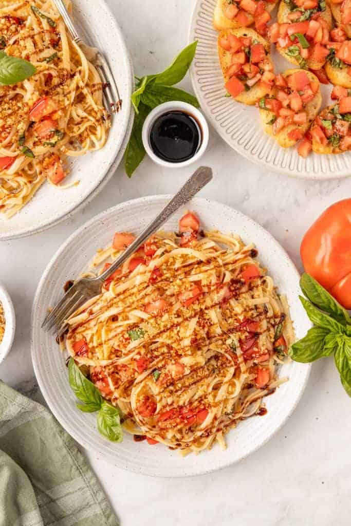 Plate of linguine pasta topped with diced tomatoes, basil, grated cheese, and balsamic glaze sits beside bruschetta, fresh basil, and a tomato—perfect for those seeking classic Italian Summer Recipes.