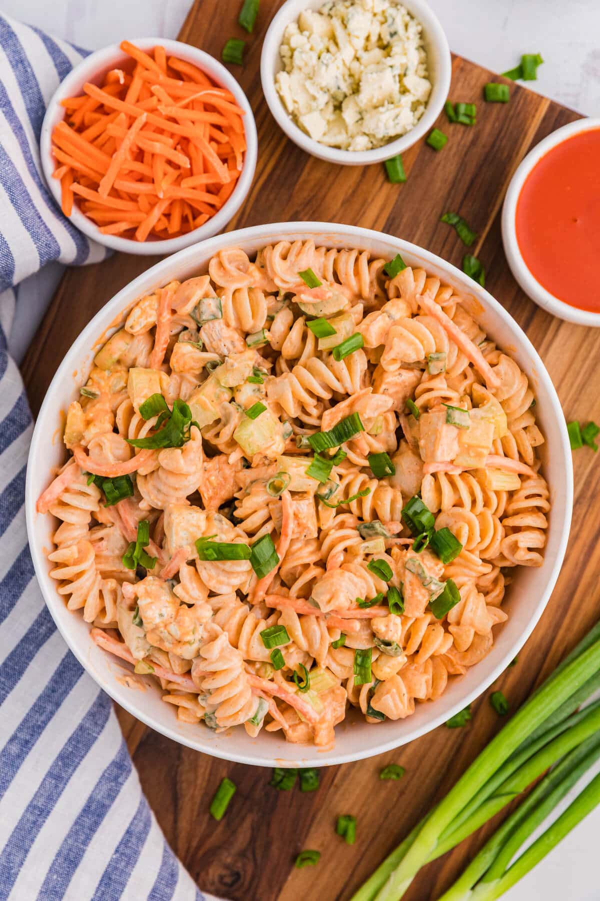 A bowl of rotini pasta salad with chopped green onions, served with shredded carrots, blue cheese, and buffalo sauce on a wooden board—perfect for summer pasta recipes.