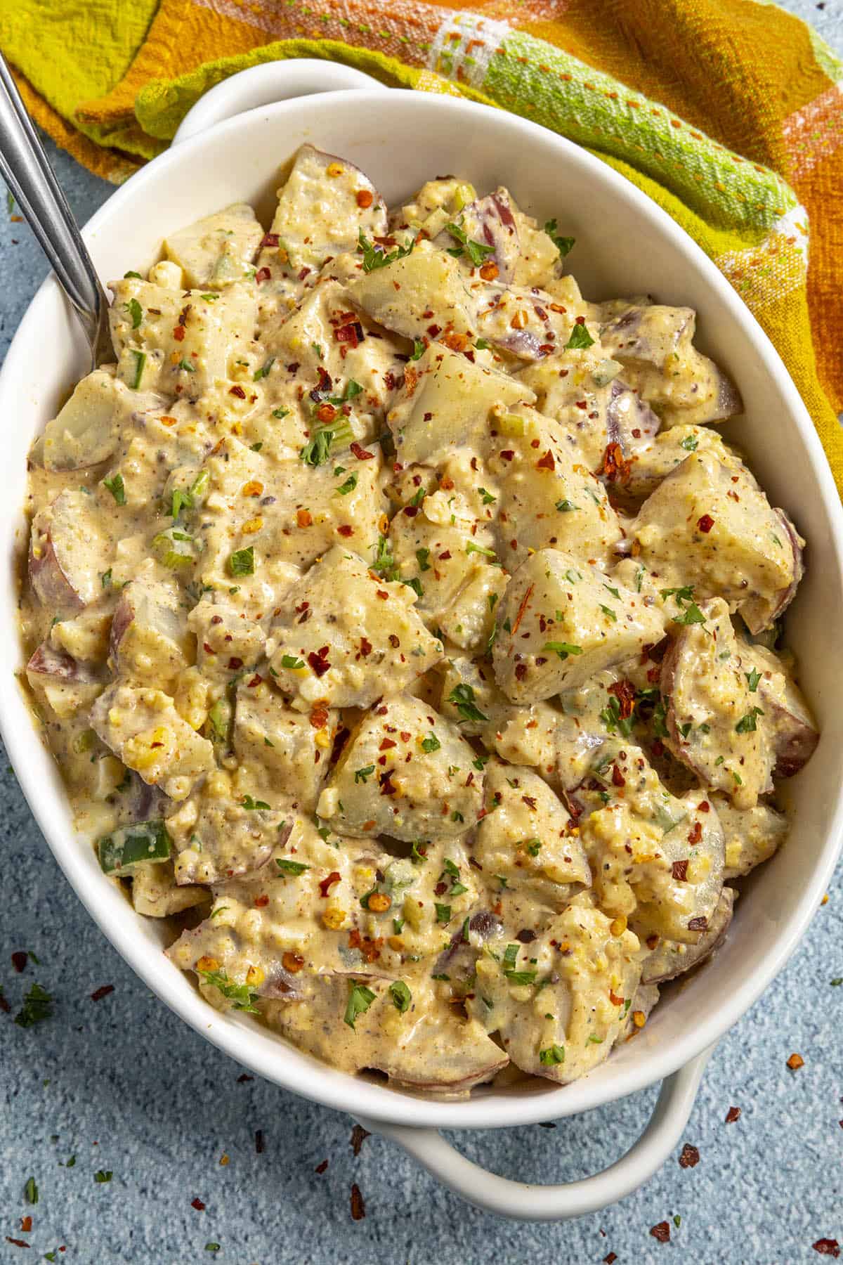 A white bowl filled with creamy potato salad, one of the perfect side dishes for any graduation party, garnished with chopped herbs and red pepper flakes, with a serving spoon inside.