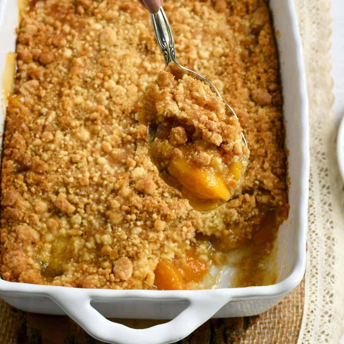 A spoon scoops out a portion of peach dessert cobbler with a crumbly topping from a white baking dish.