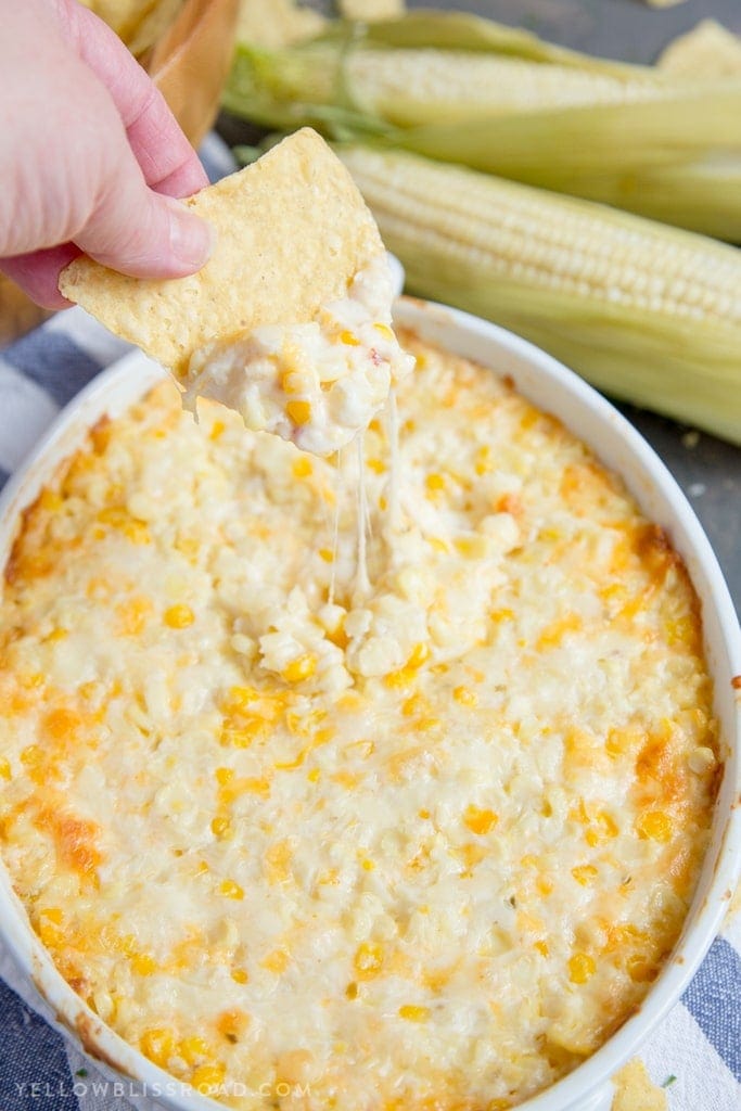 A hand dips a tortilla chip into a baked cheesy corn dip in a white oval dish, perfect for side dishes at your next graduation party, with fresh corn on the cob in the background.