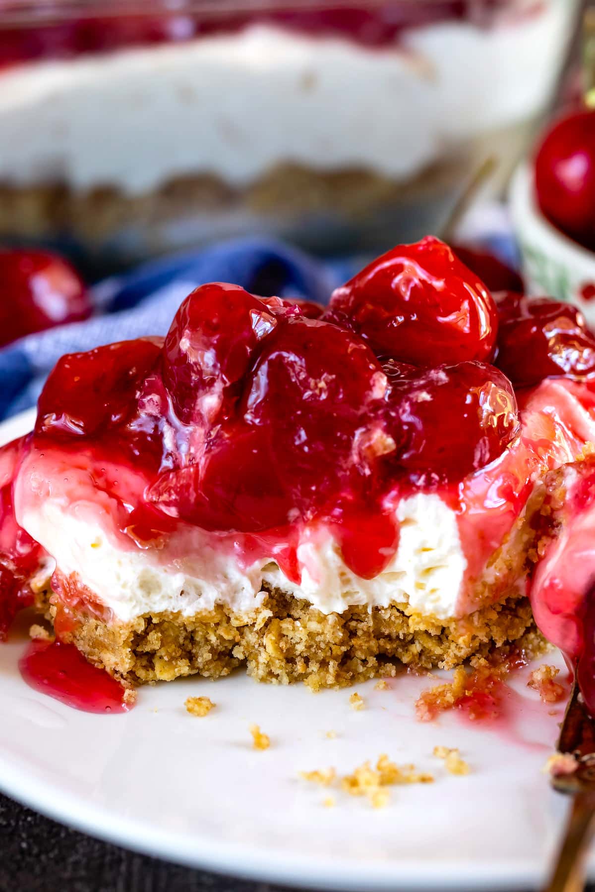A slice of cherry cheesecake with a graham cracker crust, creamy filling, and cherry topping sits on a white plate—a perfect summer dessert.