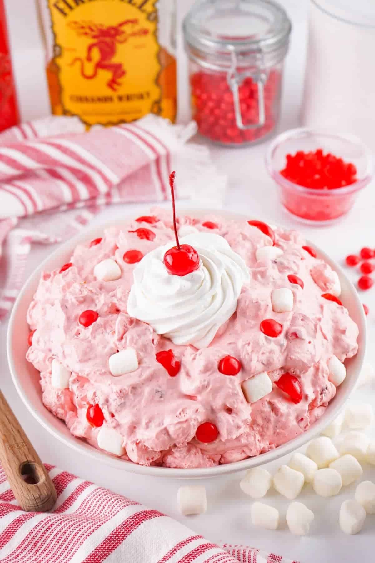 A bowl of pink marshmallow salad topped with whipped cream and a cherry, decorated with mini marshmallows and red cherries, makes a delightful summer dessert, surrounded by fresh ingredients on a white surface.