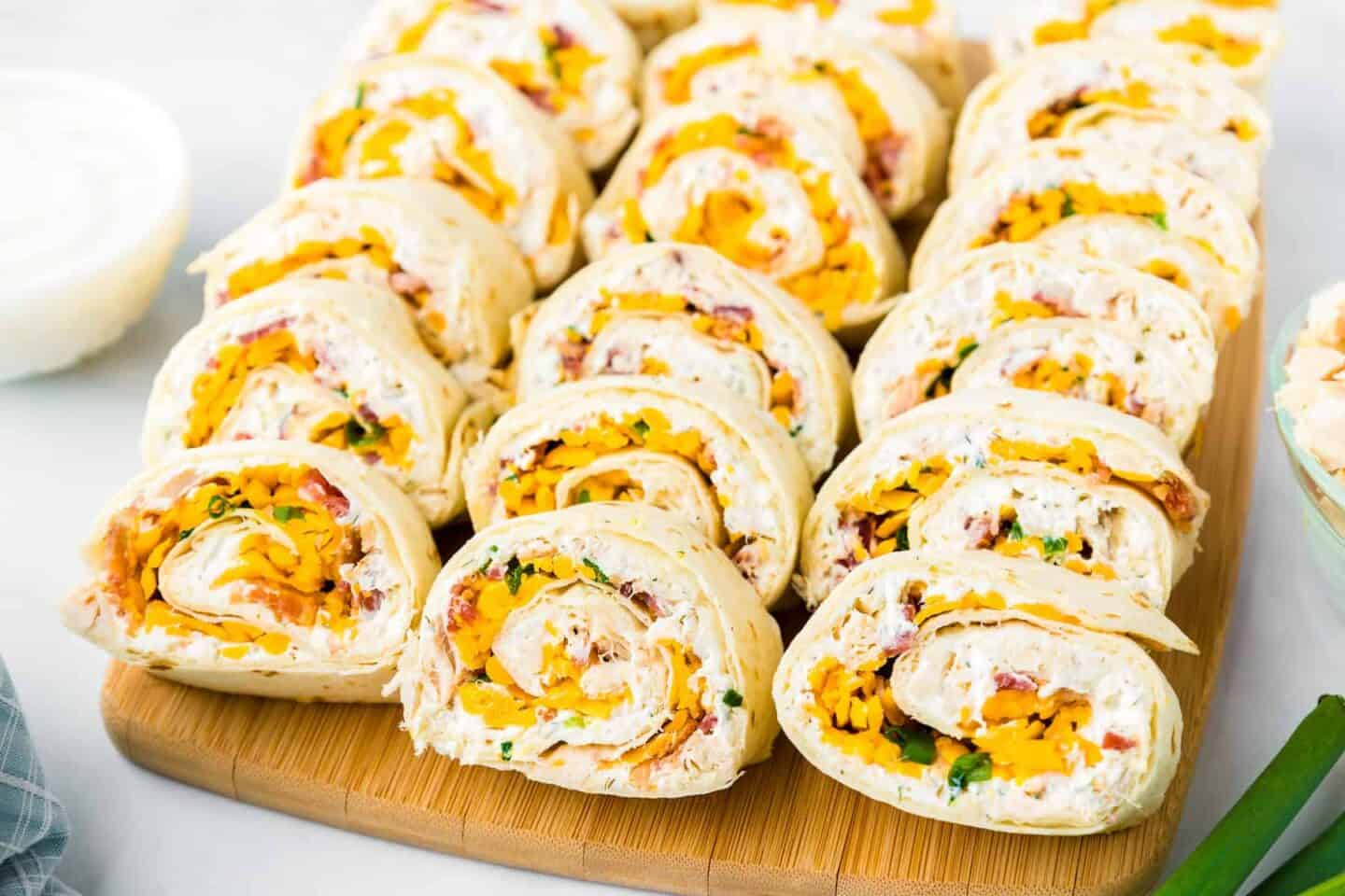 Several tortilla pinwheels filled with cream cheese, shredded cheese, bacon bits, and herbs are arranged on a wooden board—perfect for beach lunch ideas or easy picnic recipes.