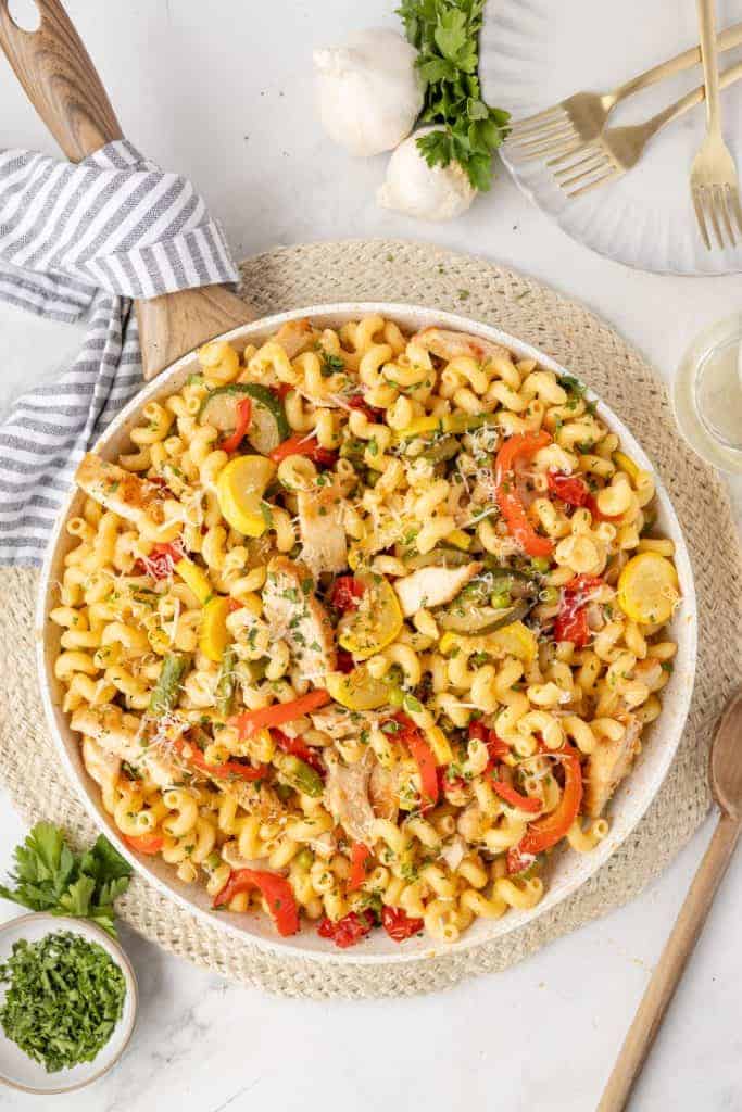 A bowl of pasta with spiral noodles, sliced chicken, red and yellow bell peppers, garnished with herbs and grated cheese—perfect for Italian Summer Recipes—on a woven placemat with utensils and fresh herbs nearby.